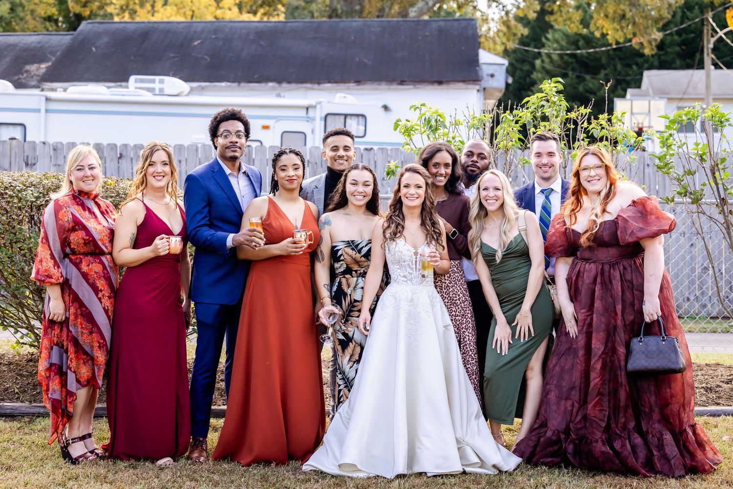 Wedding party poses outdoors in burgundy and orange dresses against natural backdrop.