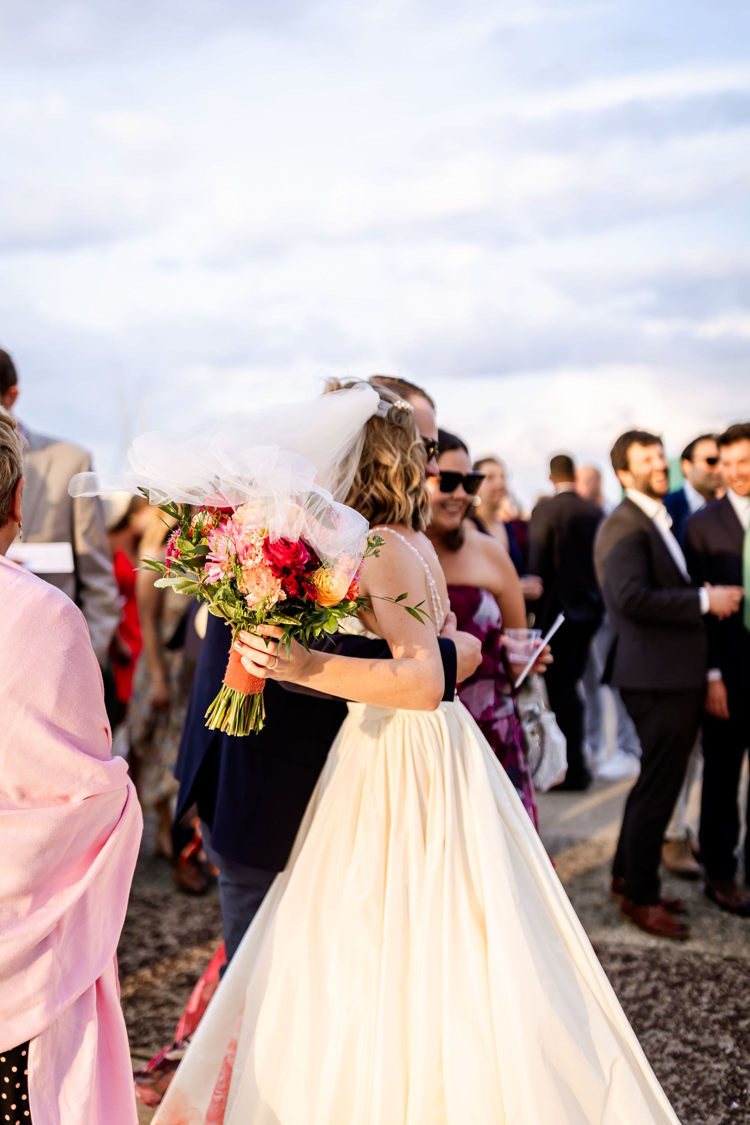 Wedding celebration on the beach with guests gathering around the bride holding a colorful bouquet.