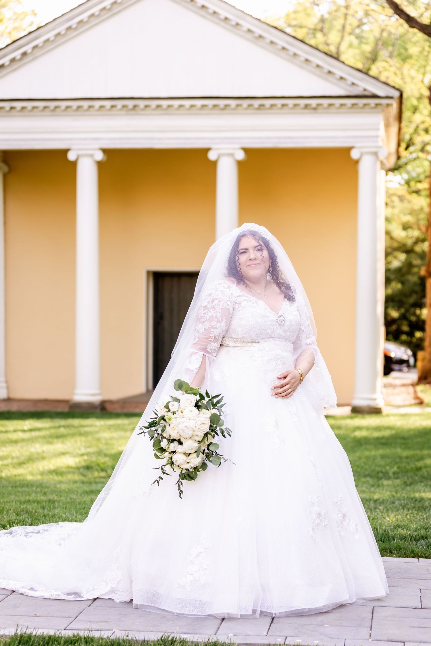 Bride in lace wedding gown and veil poses in front of classical columns and yellow building.