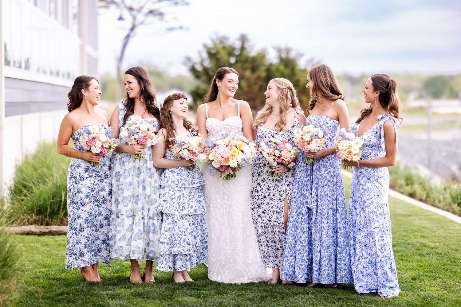 Bridesmaids wearing coordinating blue floral dresses stand together holding pink and white bouquets at an outdoor venue.