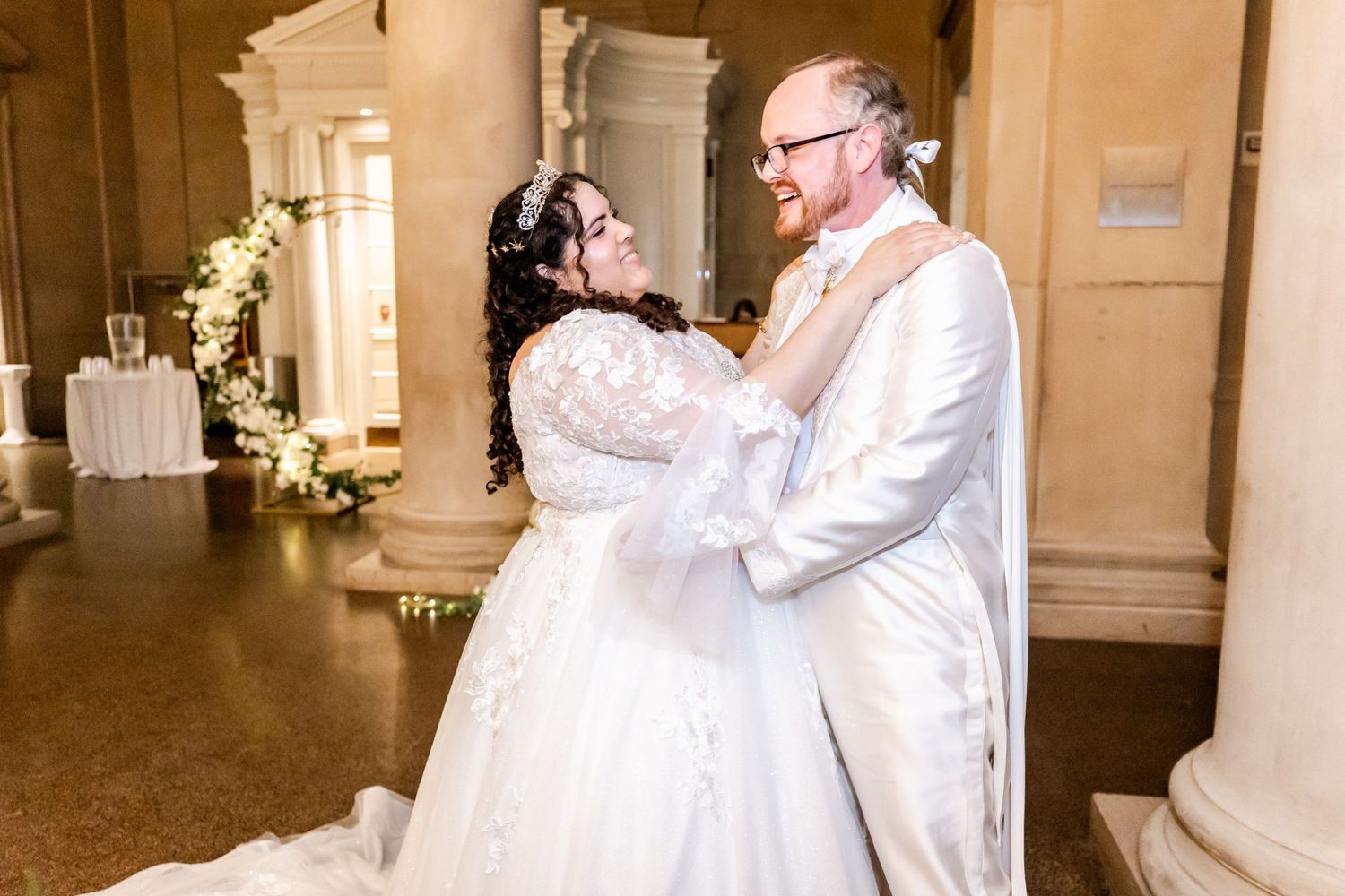 A couple shares their first dance in a grand hallway with elegant columns and floral decorations.