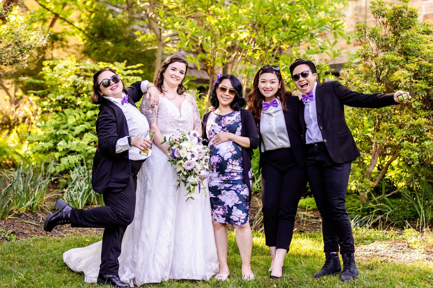 Wedding party in formal attire posing together outdoors with floral bouquets against greenery.