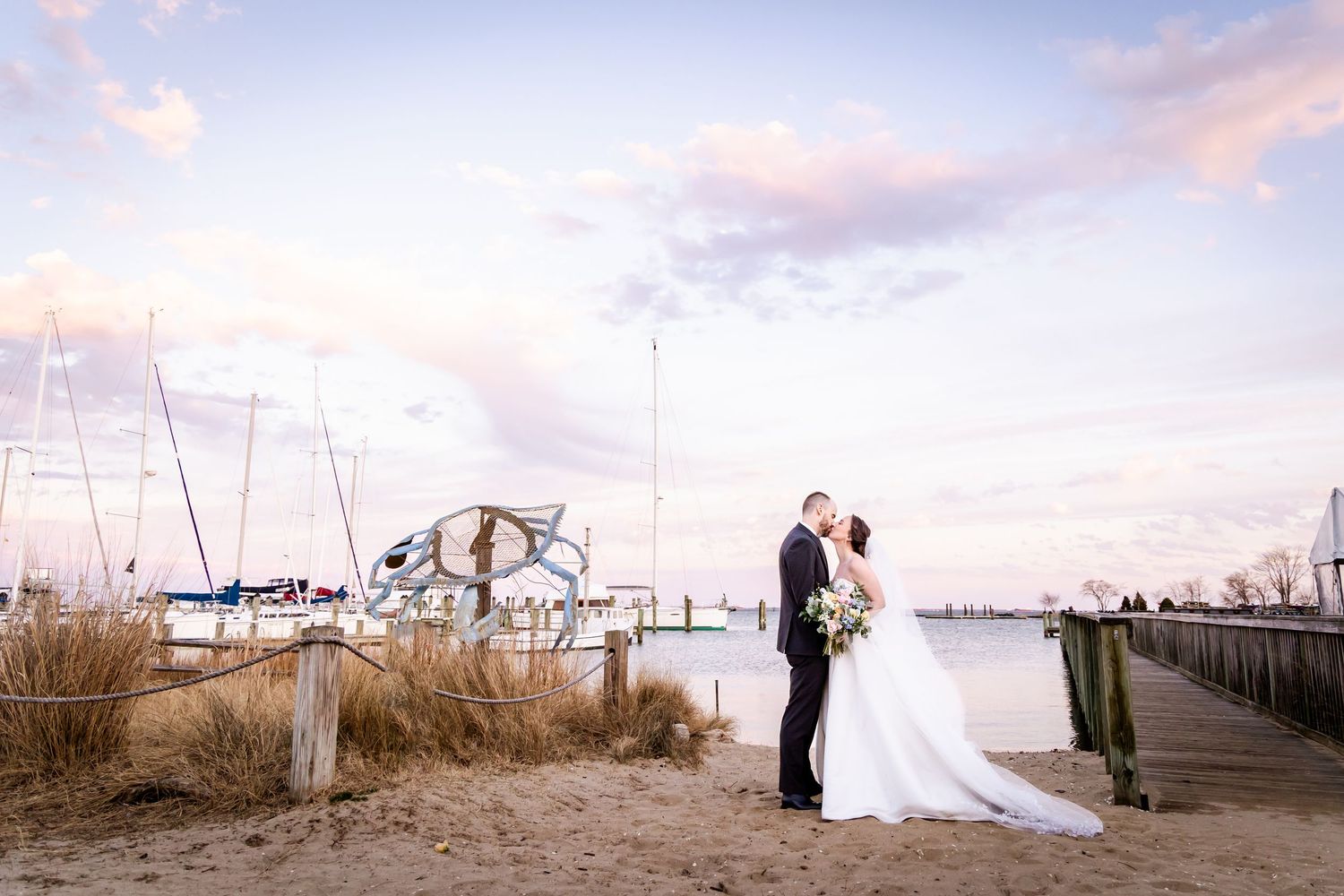 Newlyweds share a romantic moment on a marina dock at sunset with sailboats and pink clouds in background.