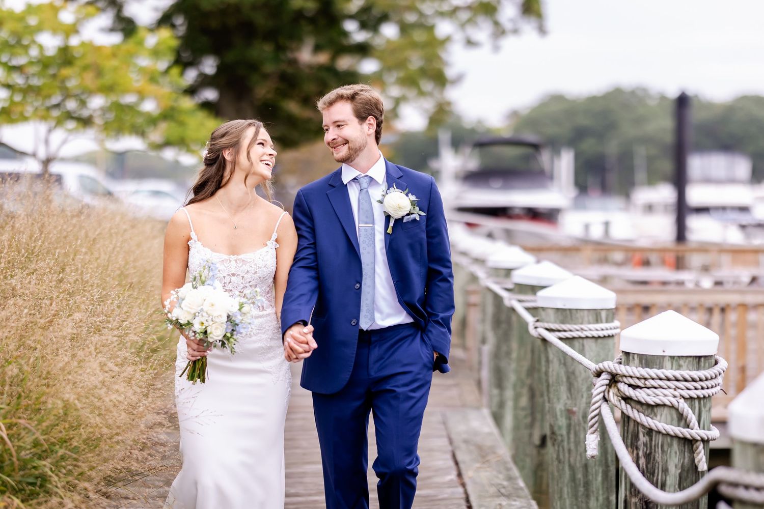 Couple in navy blue and white attire walking together on a wooden dock overlooking a marina on their wedding day.