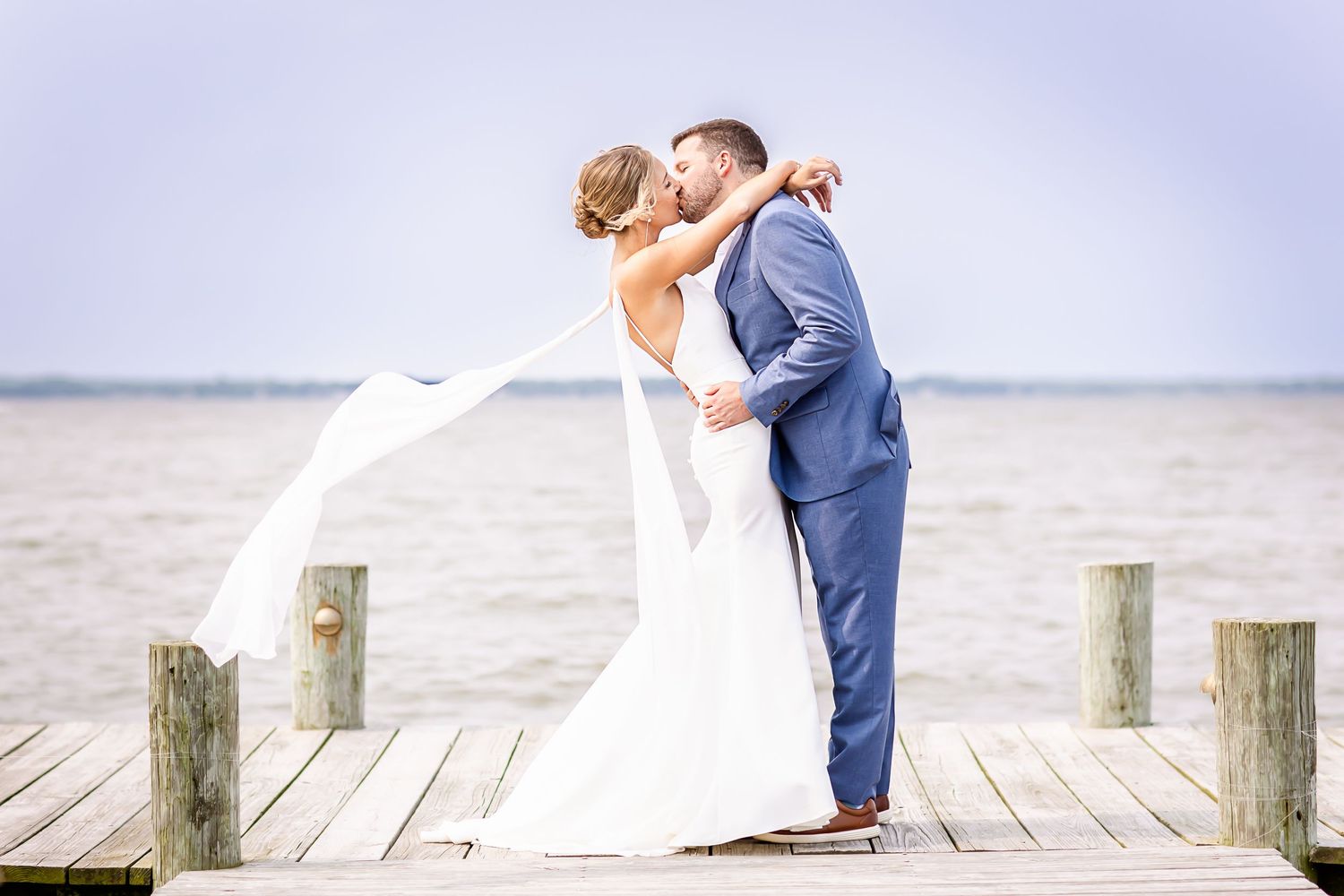 Newlyweds share a romantic moment on a wooden pier by the water as bride's veil flows in the breeze.