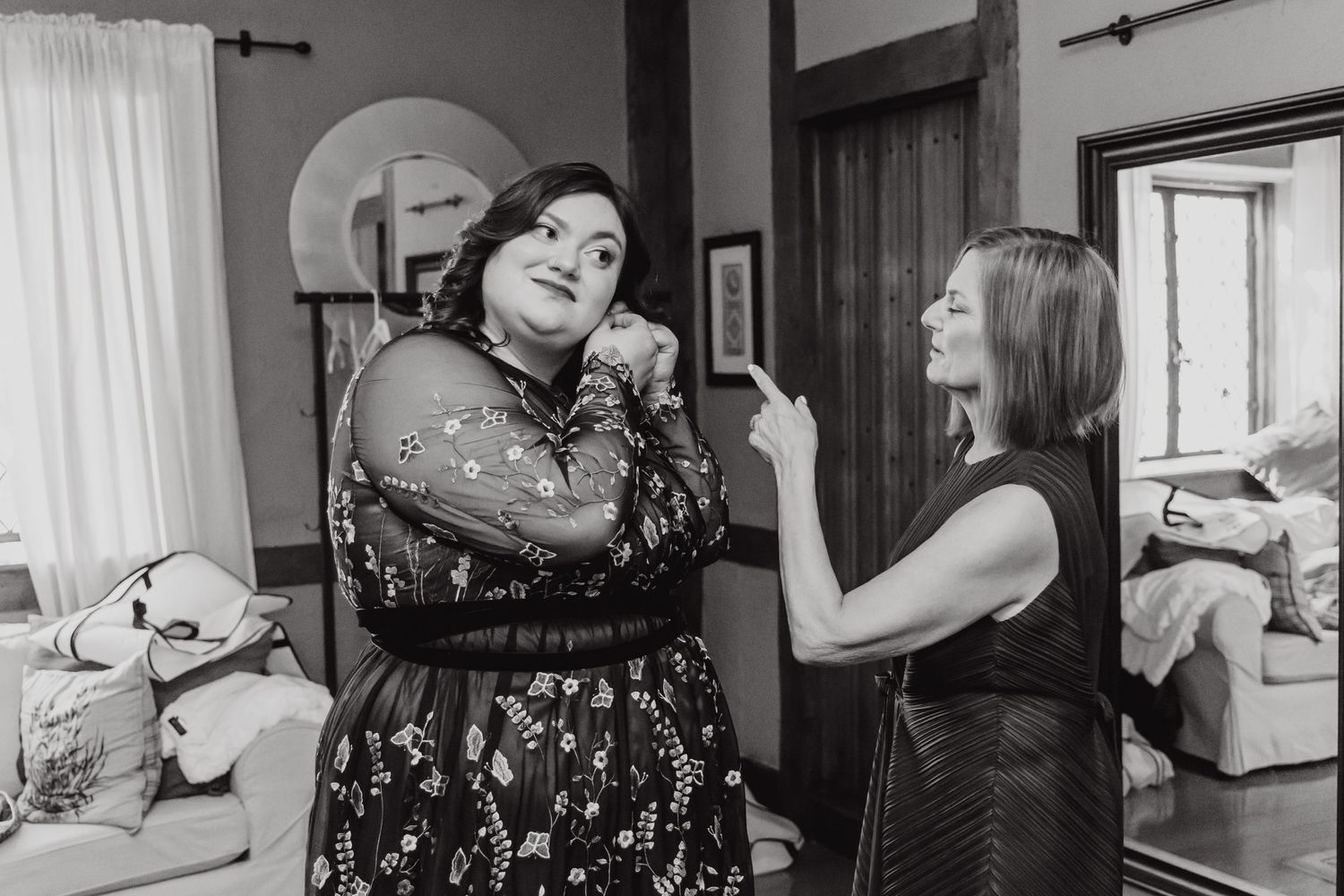Two women in dresses share an animated moment while laughing and gesturing in a living room.
