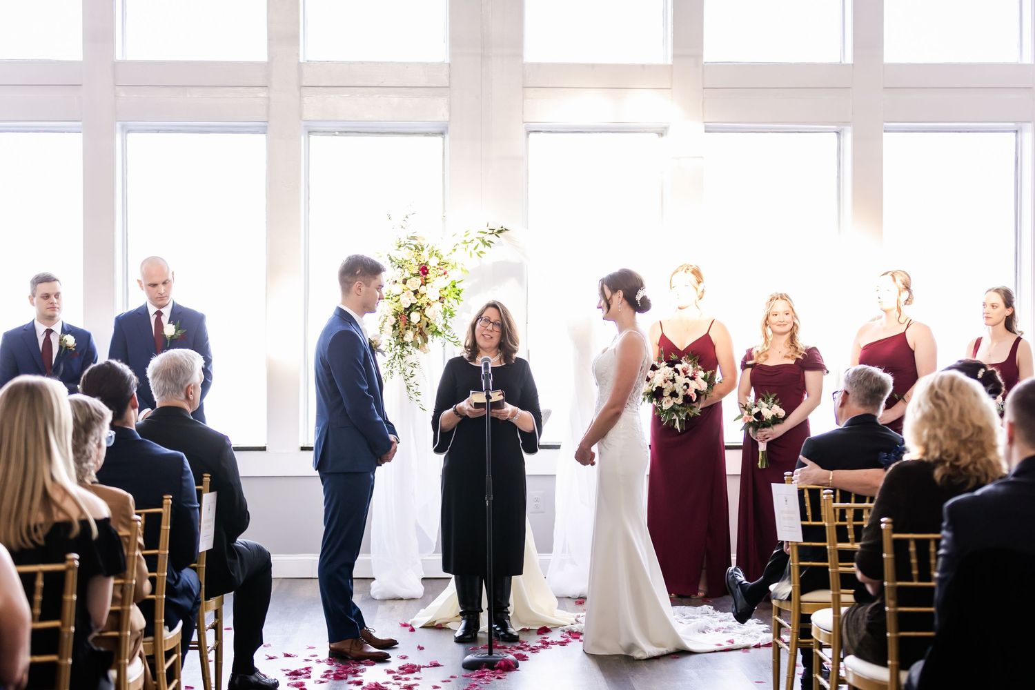 Wedding ceremony guests seated in bright indoor venue with large windows.
