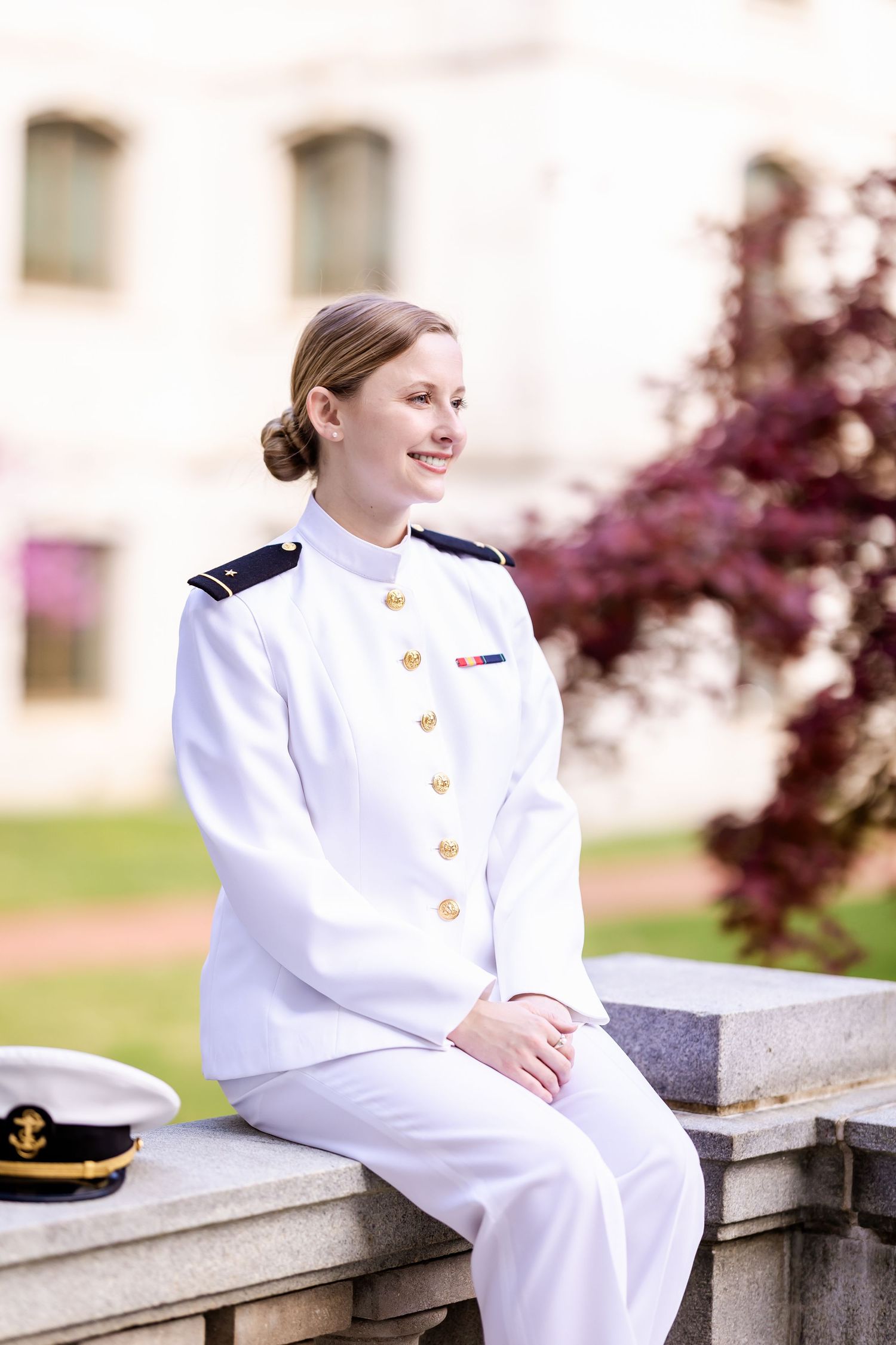 A person in formal white naval attire sits on a stone ledge with trees and buildings visible in the background.
