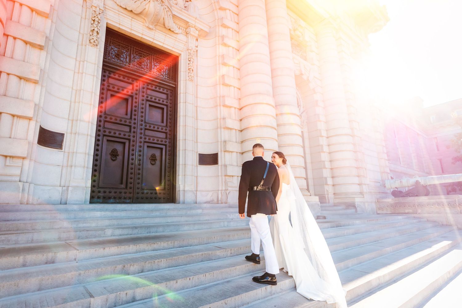 A couple in wedding attire stands on stone steps in front of large wooden doors, with sunlight creating a dreamy glow.