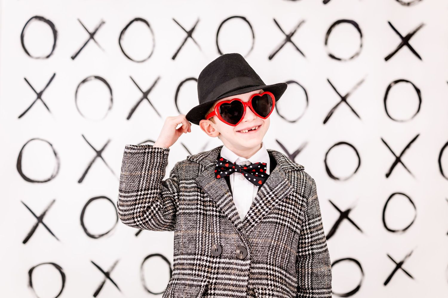 A stylishly dressed figure in plaid jacket and bowtie poses against a wall decorated with X's and O's pattern.