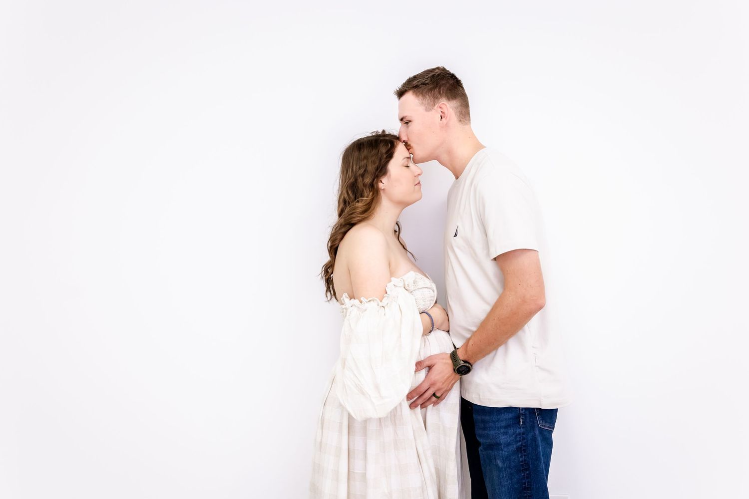 An intimate maternity portrait of a couple in white sharing a tender moment against a bright backdrop.