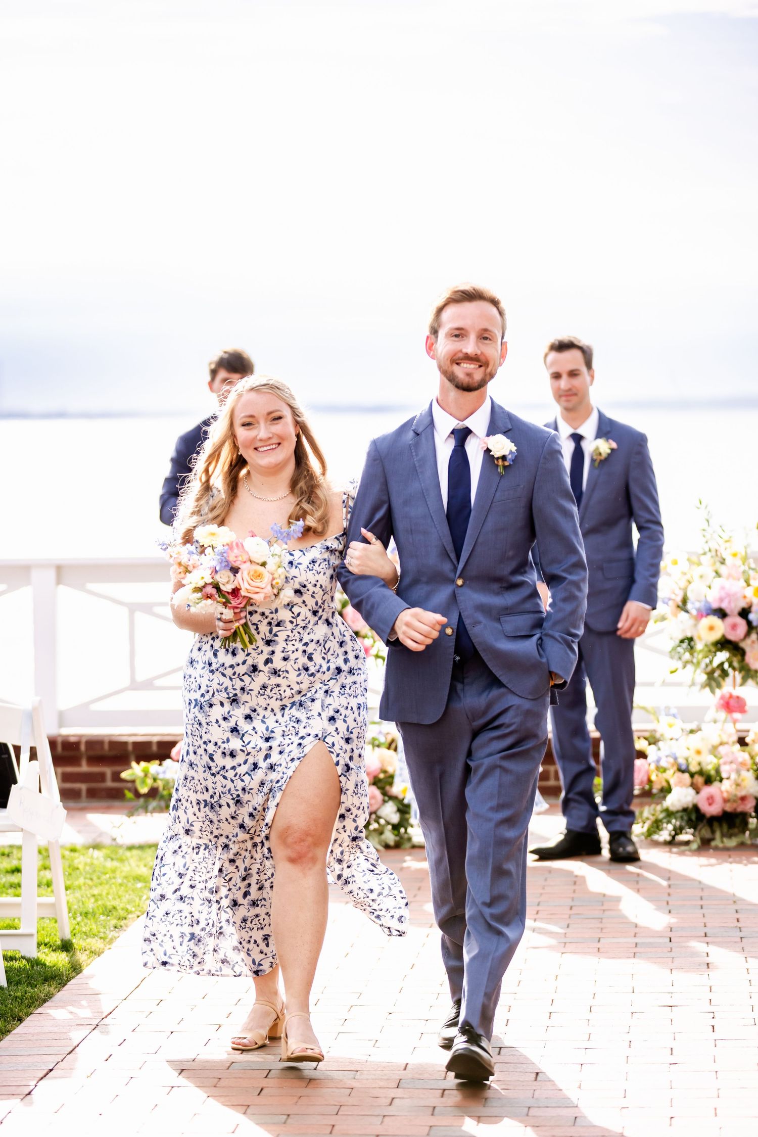 Wedding party walking along brick pathway in navy suits and floral print dresses.