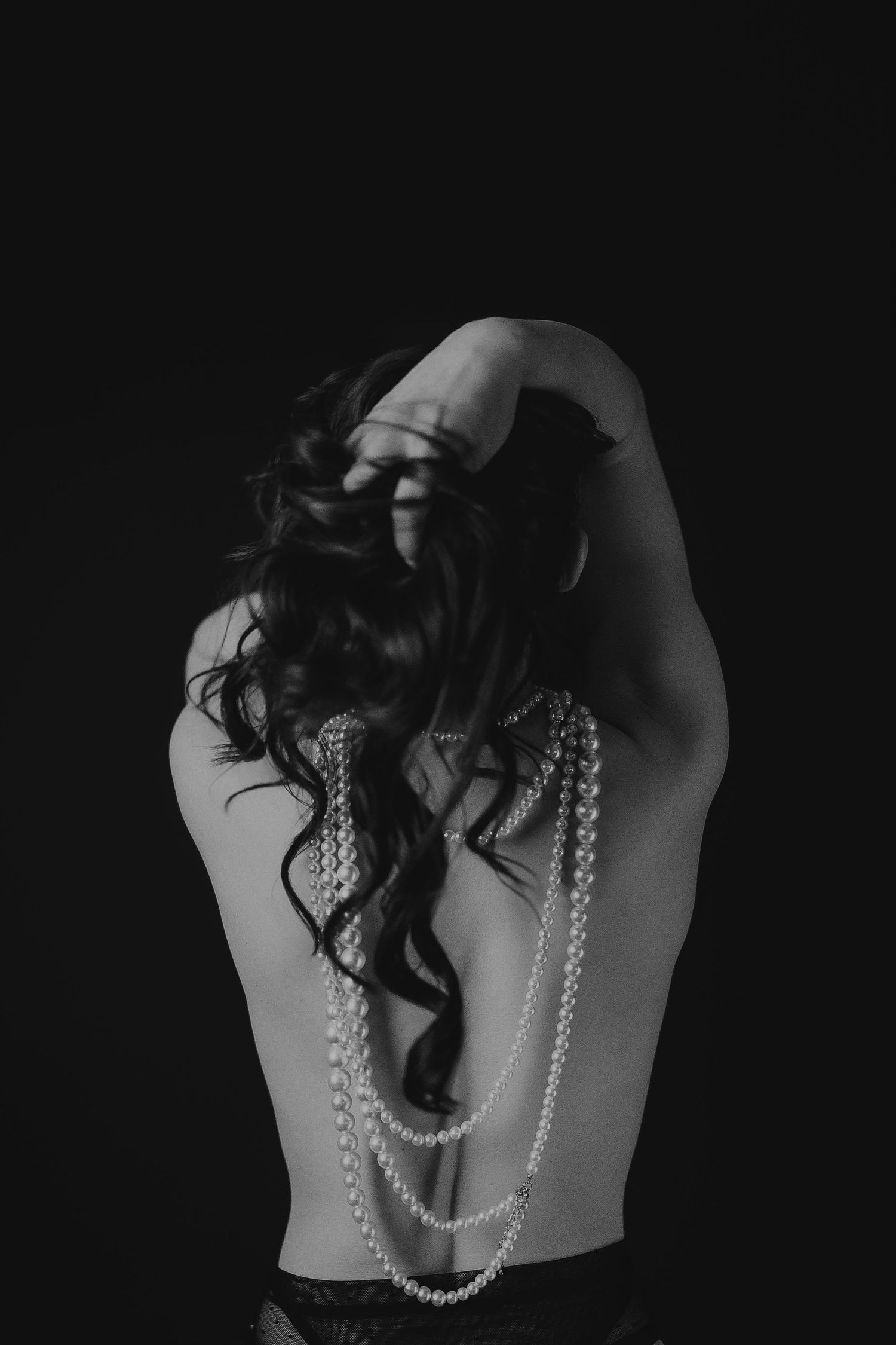 Artistic black and white photo of a bare back with draped pearl necklaces and dark curly hair against black background.