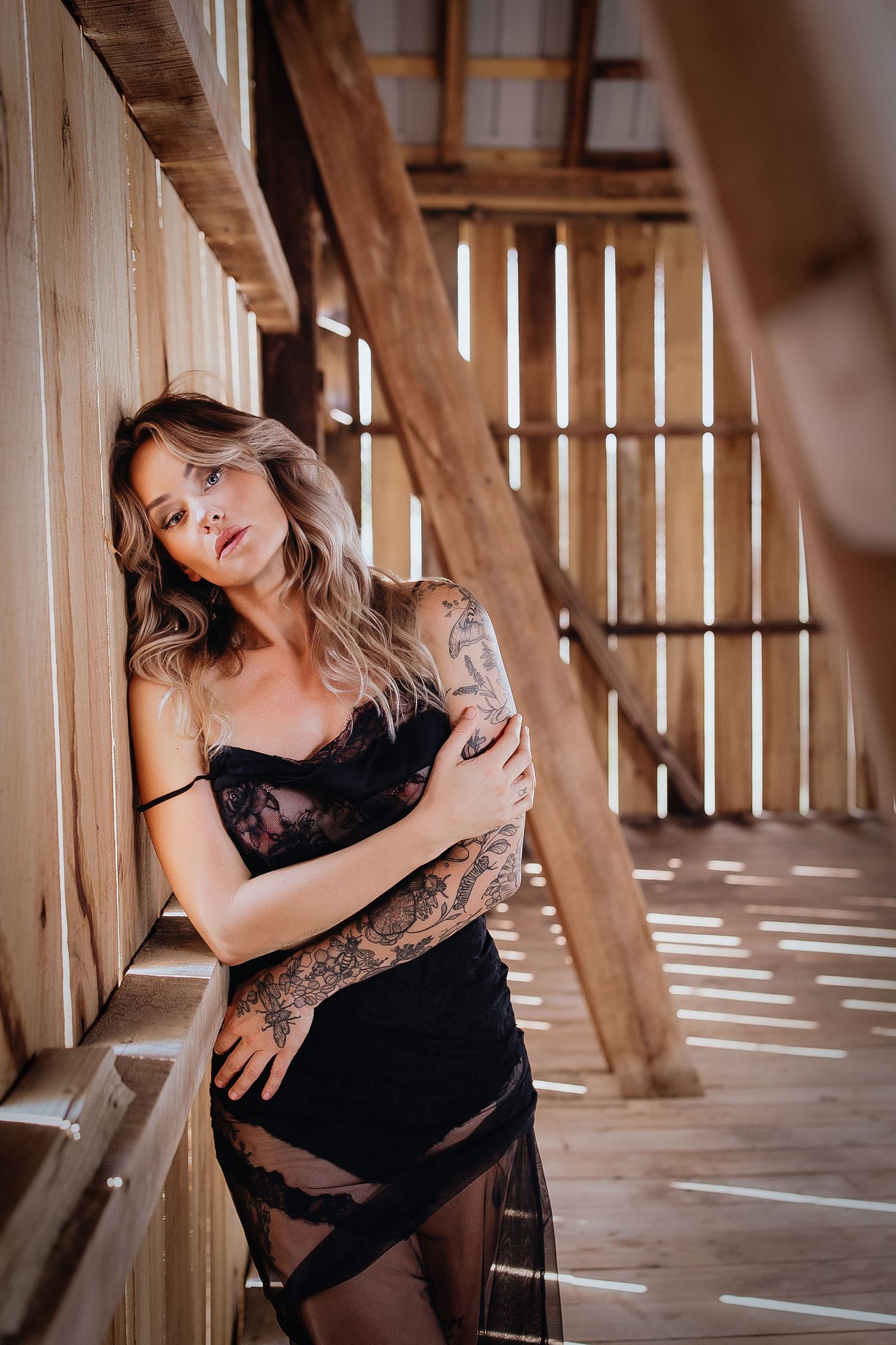Woman in black lace lingerie with tattoos leaning against wooden barn wall in moody boudoir portrait.