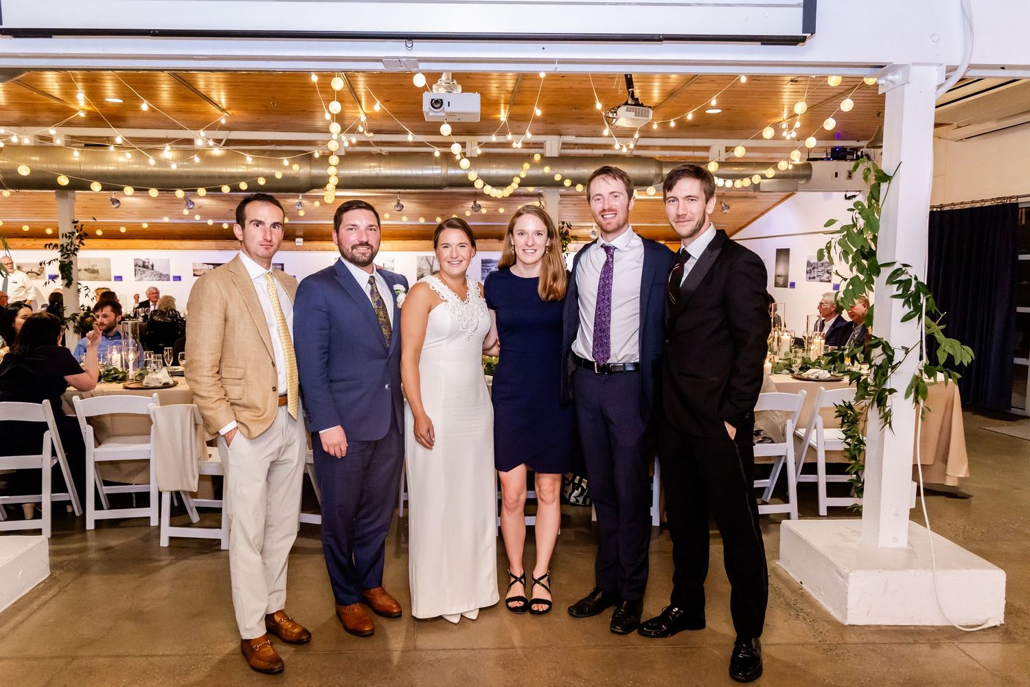 Group of six elegantly dressed guests smiling together at a wedding reception venue with string lights.