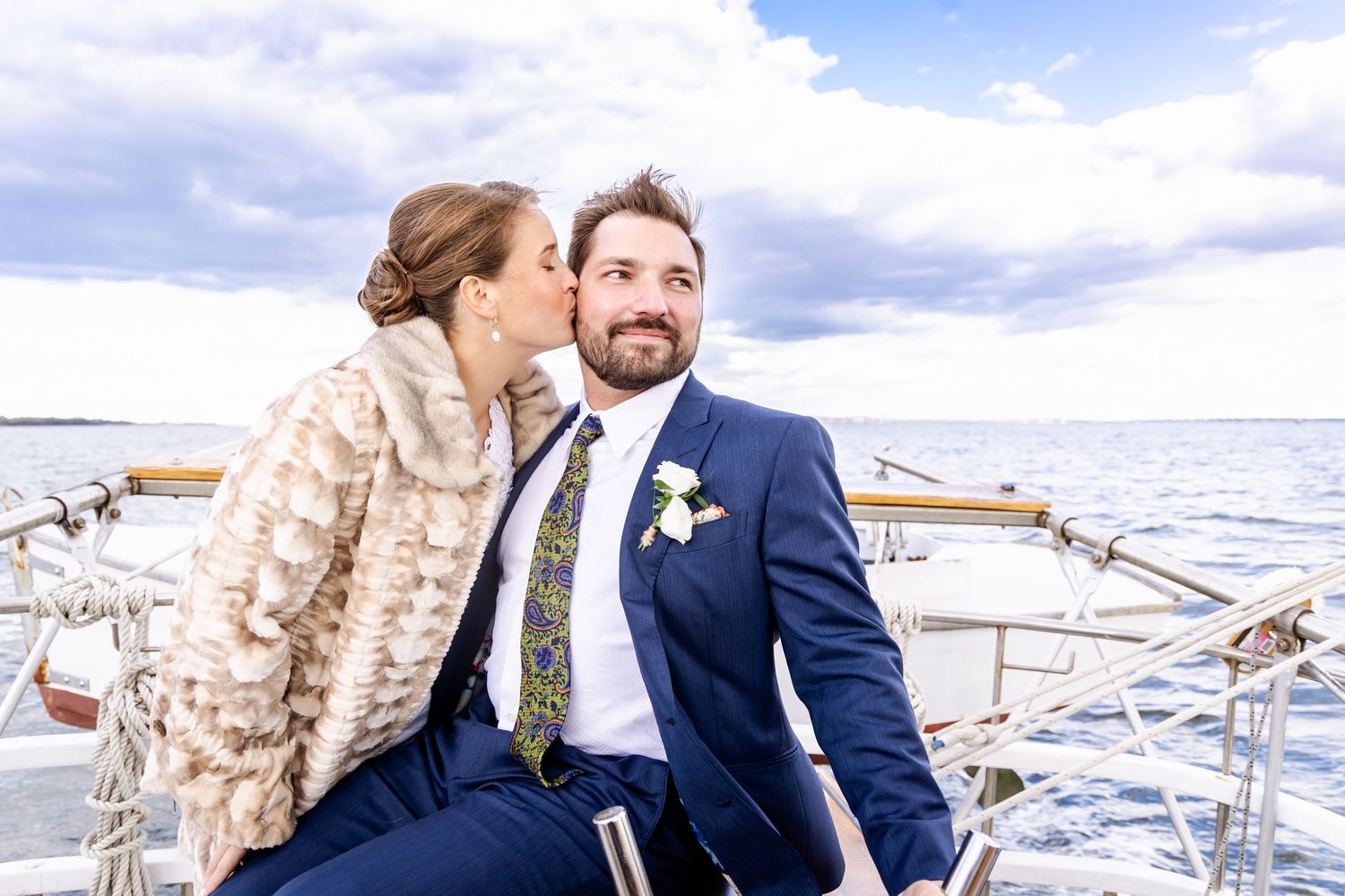 Bride kissing groom on cheek aboard sailboat on their wedding day, groom in navy suit with floral tie.