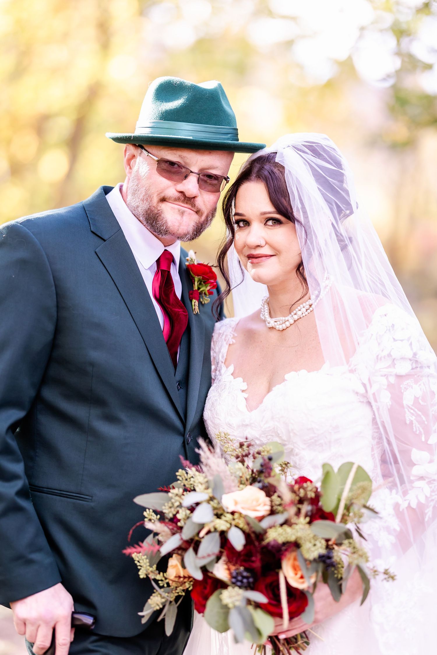 A wedding couple poses outdoors in autumn with the groom wearing a teal hat and red tie.