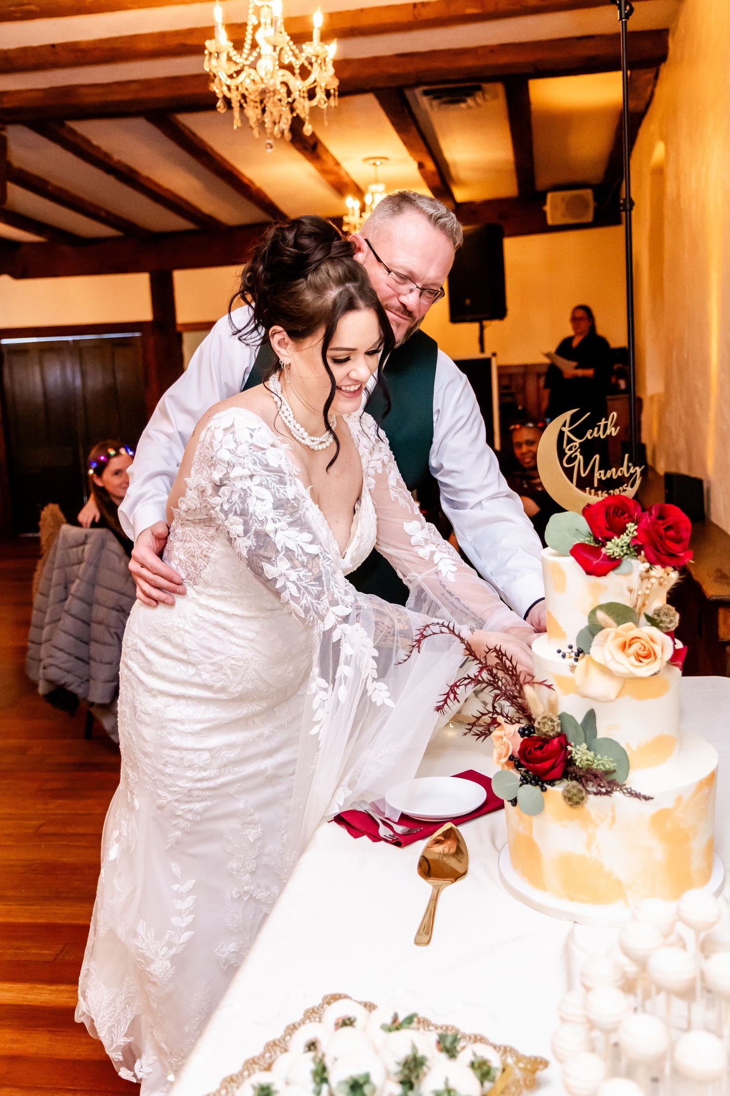 Couple cutting their wedding cake together at reception with elegant chandelier lighting and floral decorations.