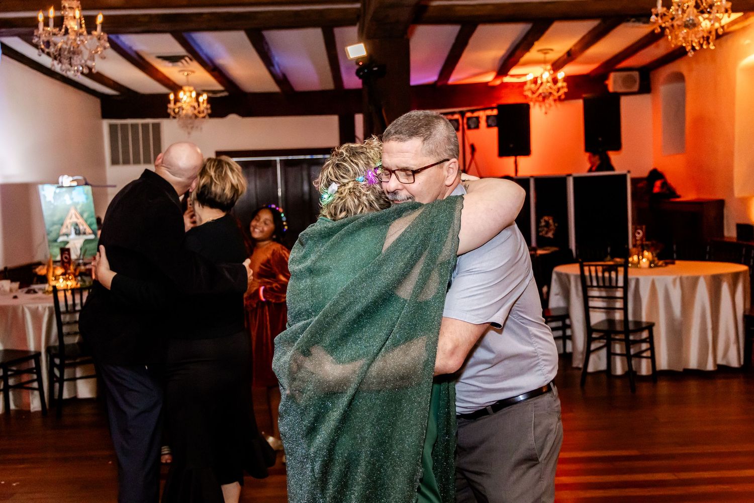 Couples dancing at a wedding reception in a rustic venue with exposed wooden beams and warm orange lighting.