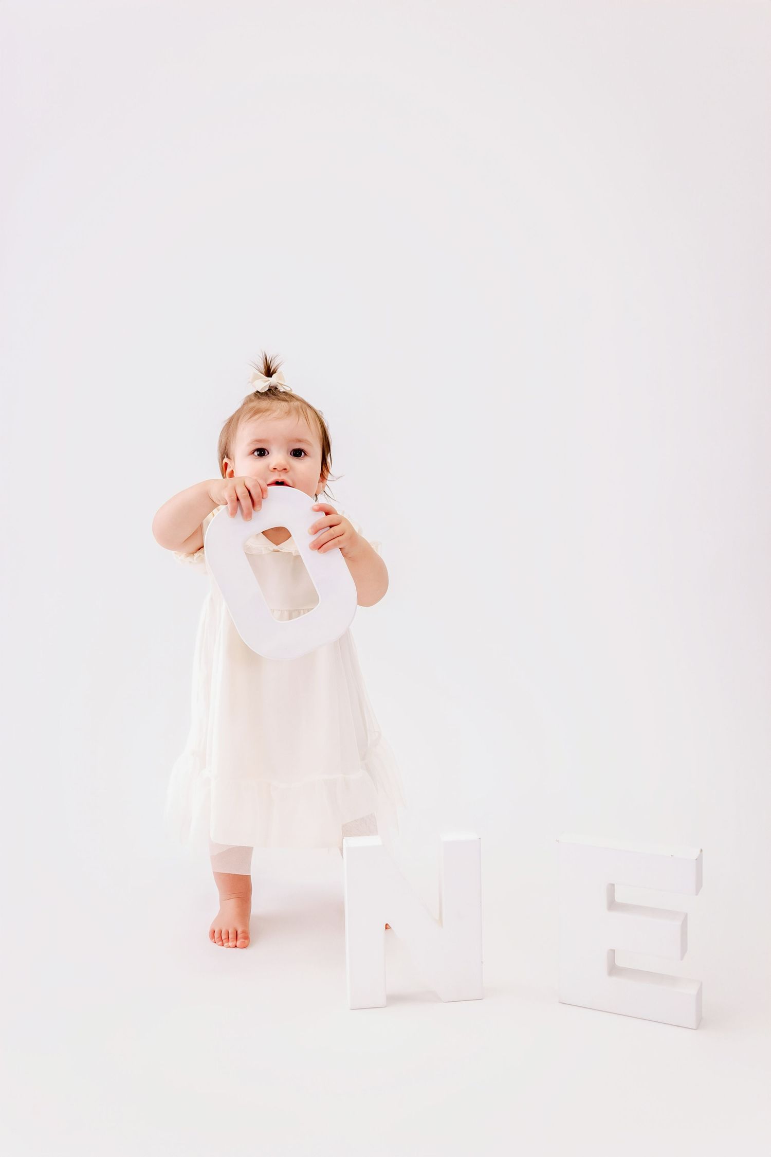 Small child in white dress standing on white background with decorative letters on the floor.