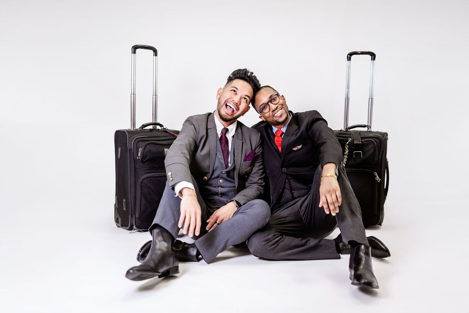 Two people in business attire sit on the floor with luggage, laughing and leaning on each other against white background.