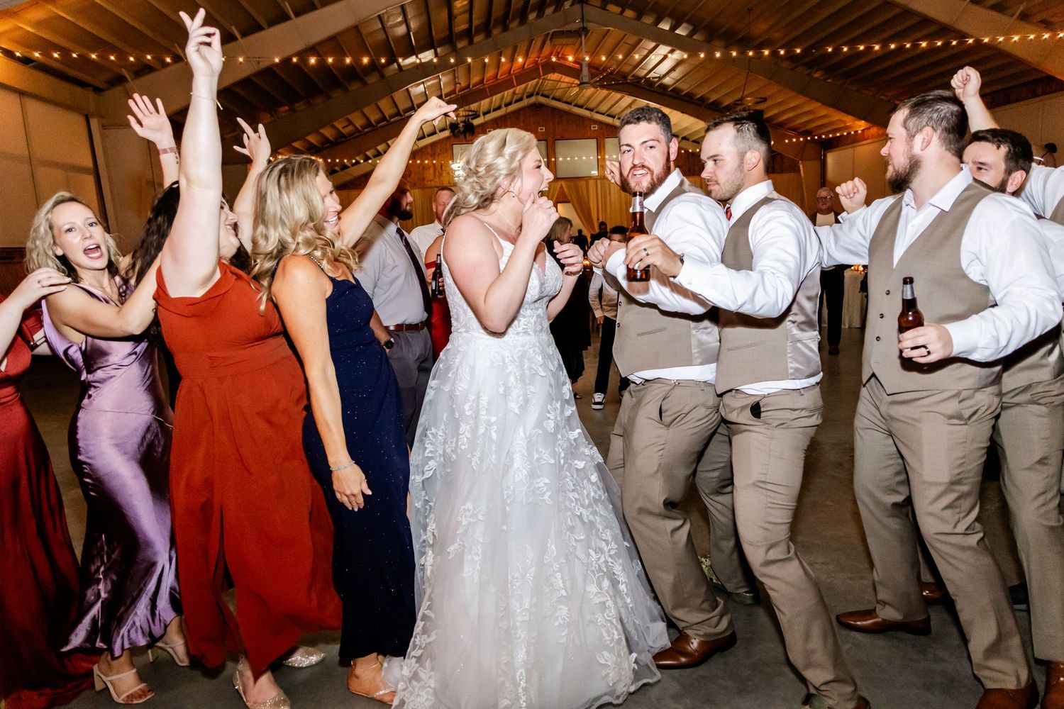Wedding reception guests dance and celebrate together on a rustic barn dance floor decorated with string lights.