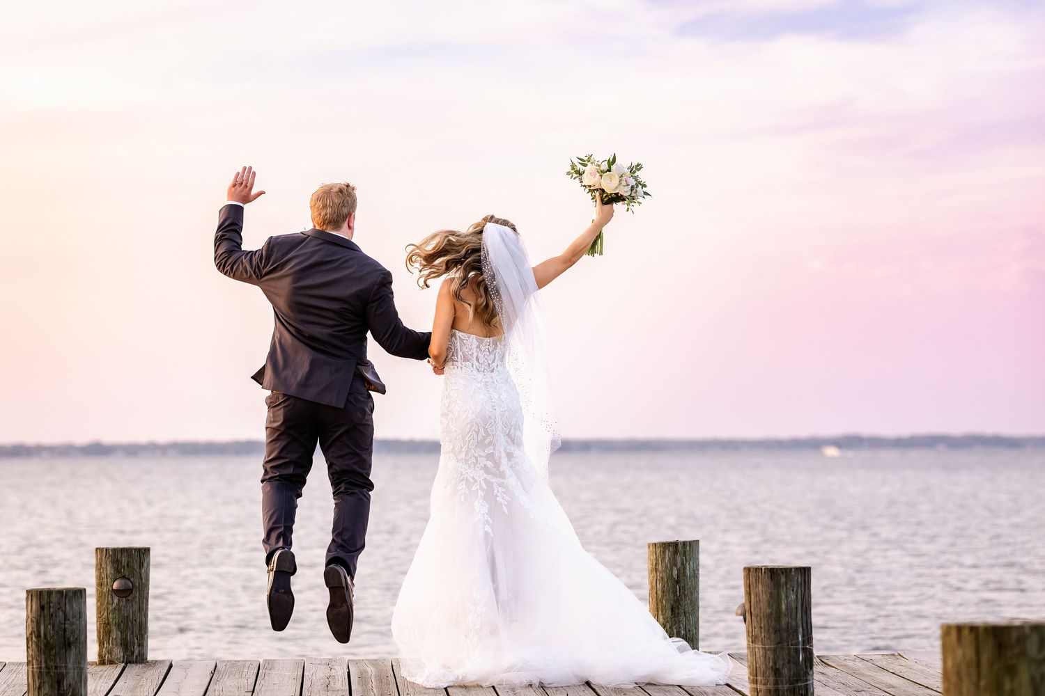 Couple in wedding attire jumps joyfully on a wooden pier at sunset over calm waters.