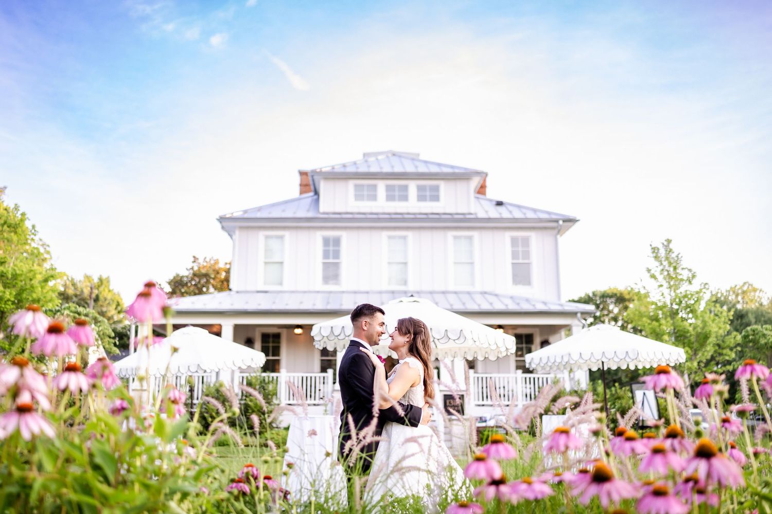 White colonial style house with pink wildflowers blooming in the foreground garden.
