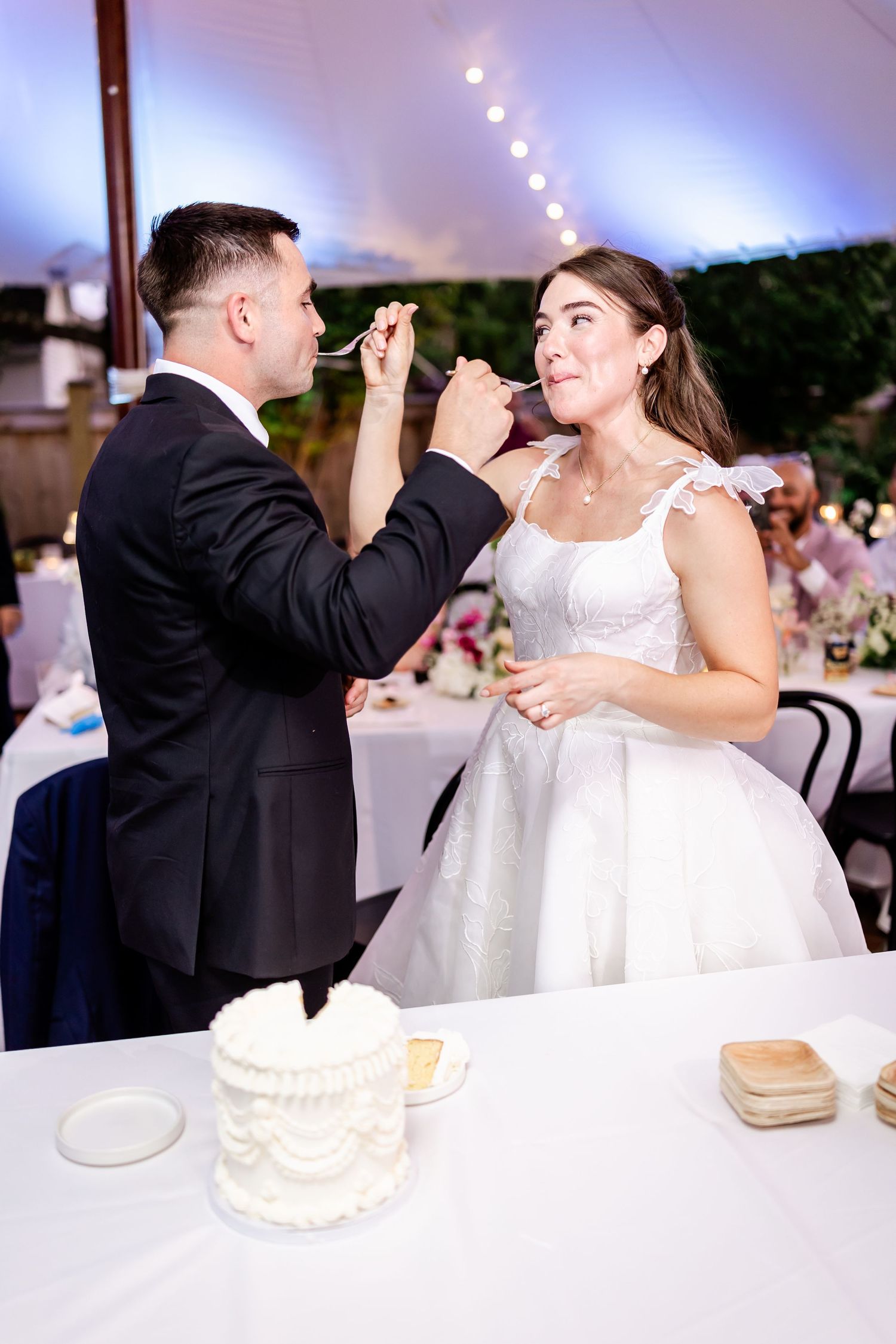 A couple shares their first bite of wedding cake together at an elegant reception under string lights.