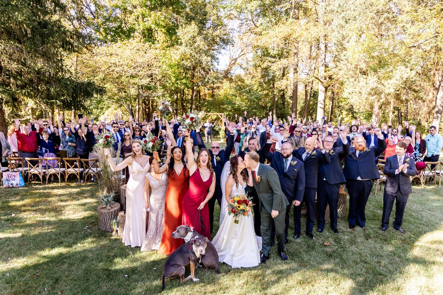 Large wedding group photo with guests gathered outdoors in natural woodland setting.