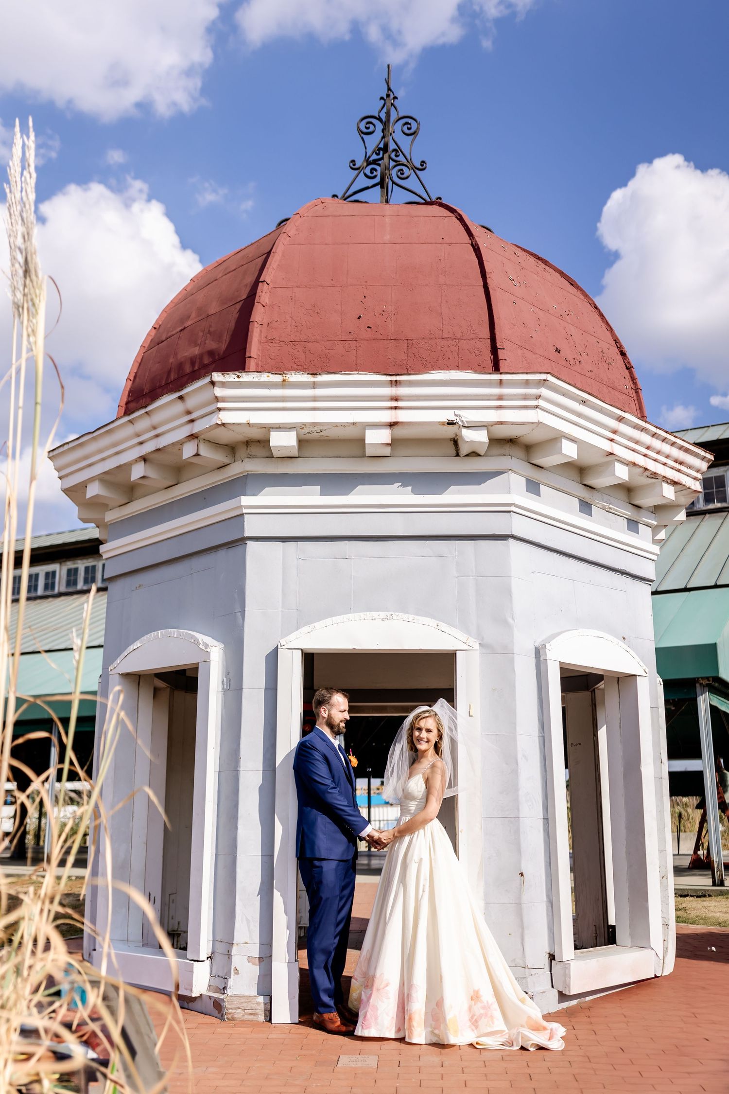 A couple stands in an ornate gazebo with a red domed roof and white columns on their wedding day.