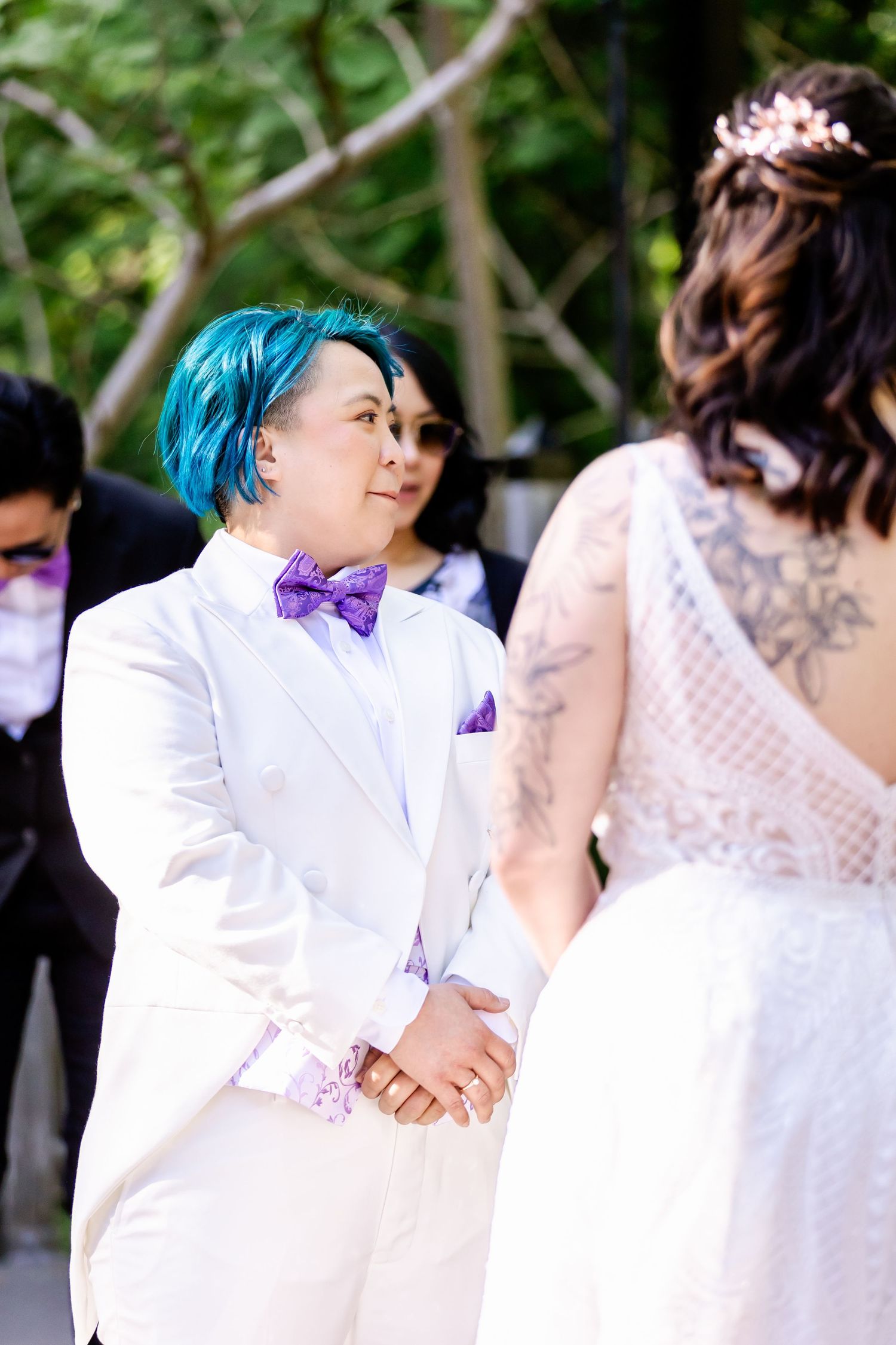 A wedding ceremony participant in a white suit and purple bow tie with vibrant blue hair.