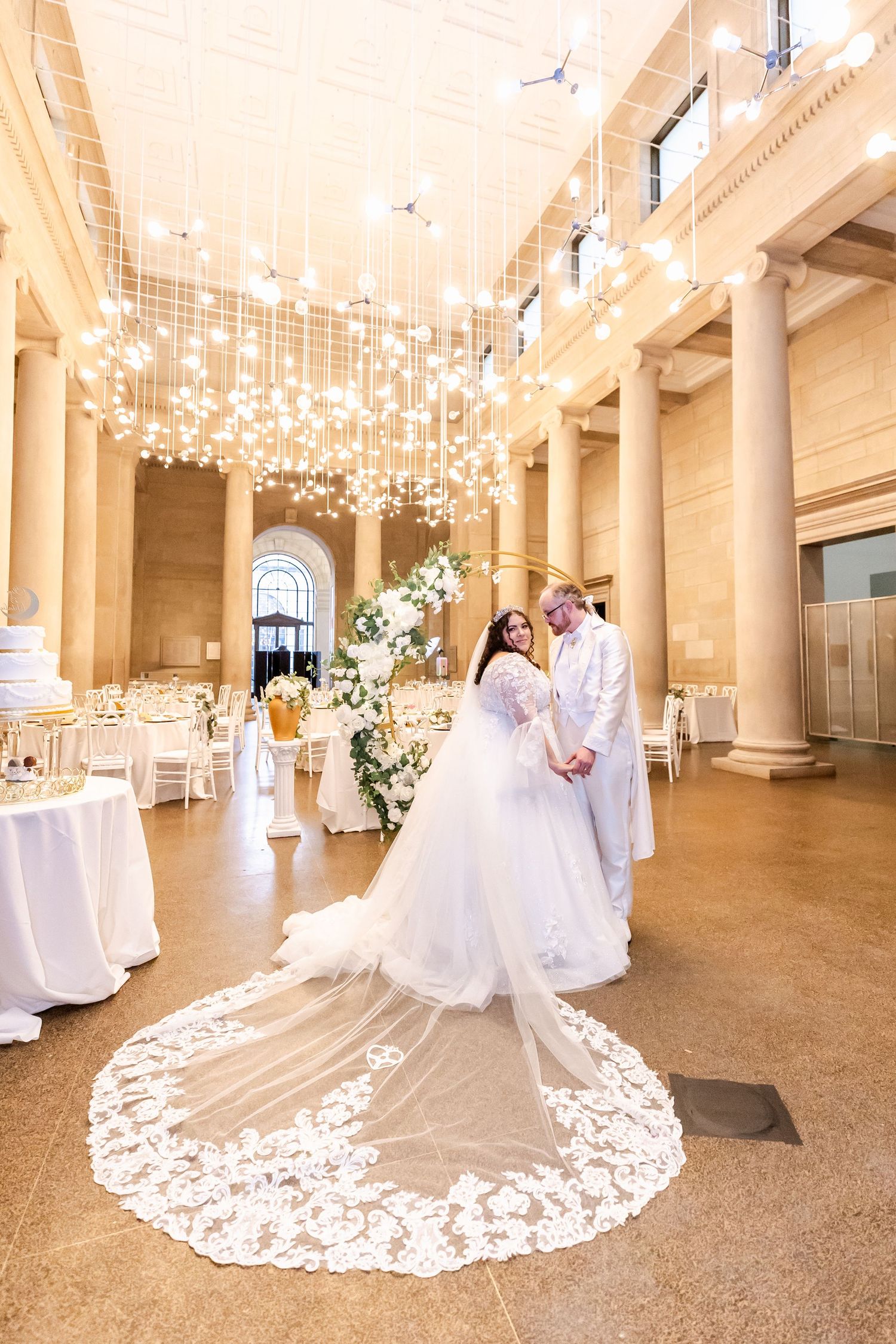 Wedding couple poses in elegant venue with hanging crystal lights and intricate lace dress train.