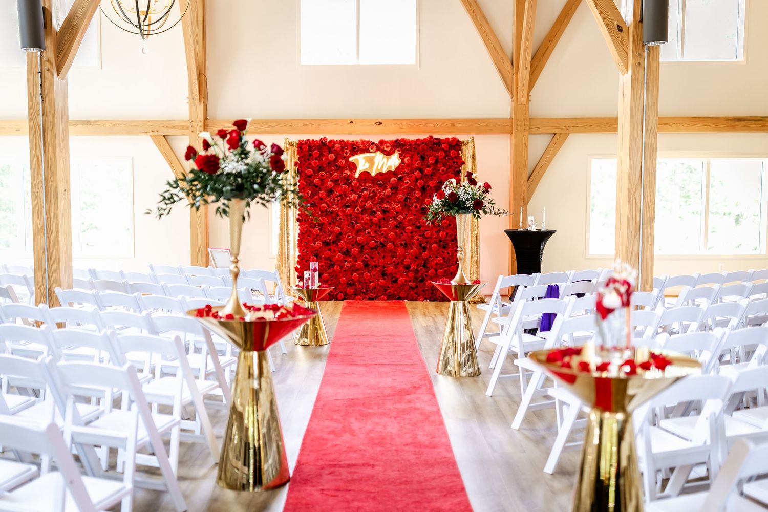 Wedding ceremony setup with red rose wall backdrop and white chairs along red carpet aisle.