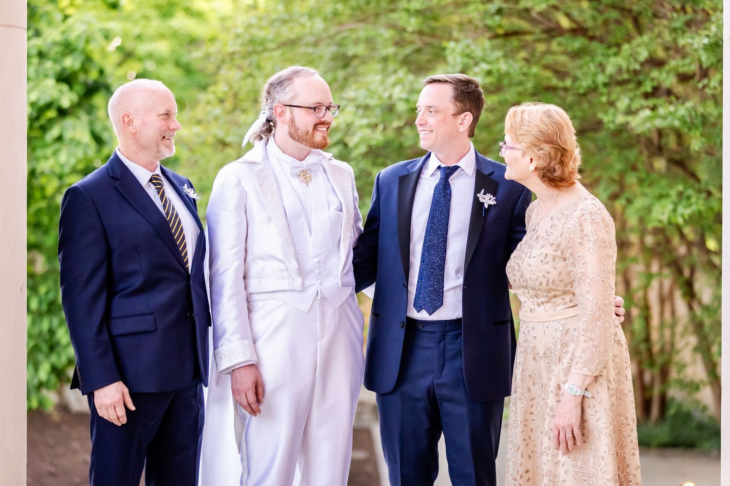 Four people in formal wedding attire share a candid moment against an outdoor backdrop.