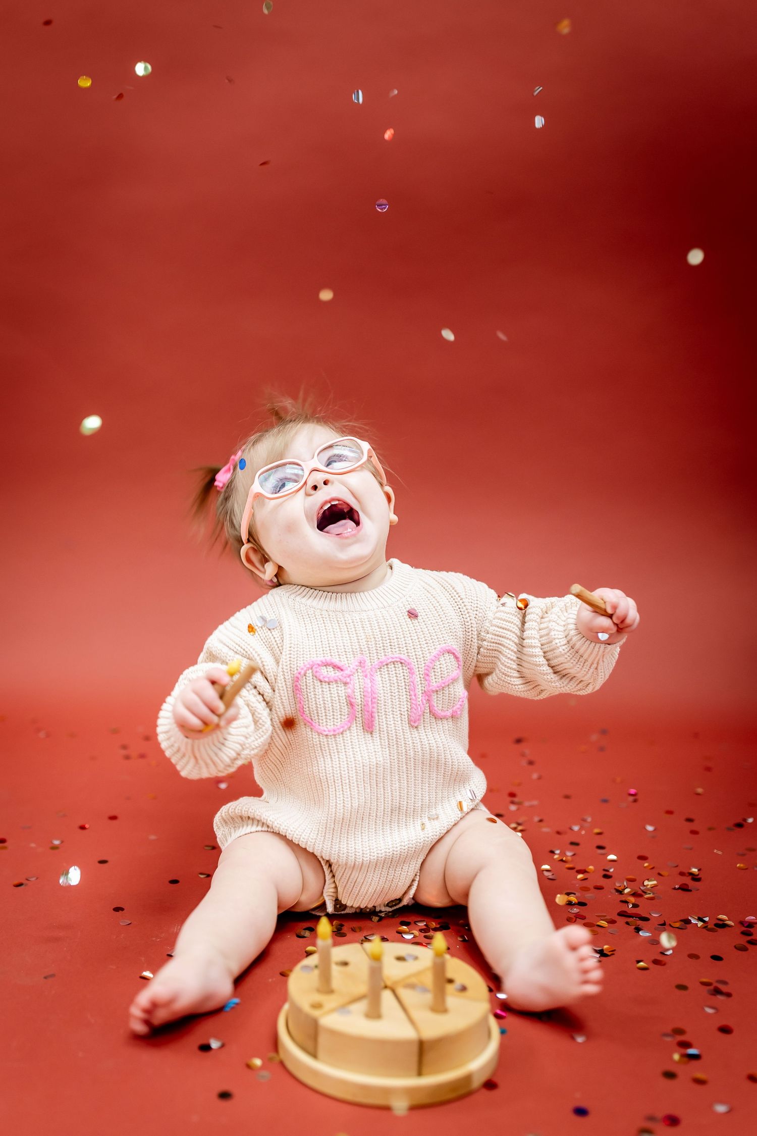 A baby in knit sweater laughs with confetti falling on red background.