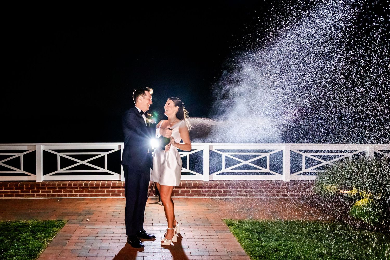 A couple stands on a patio at night as water sprays behind them creating a magical effect in the darkness.