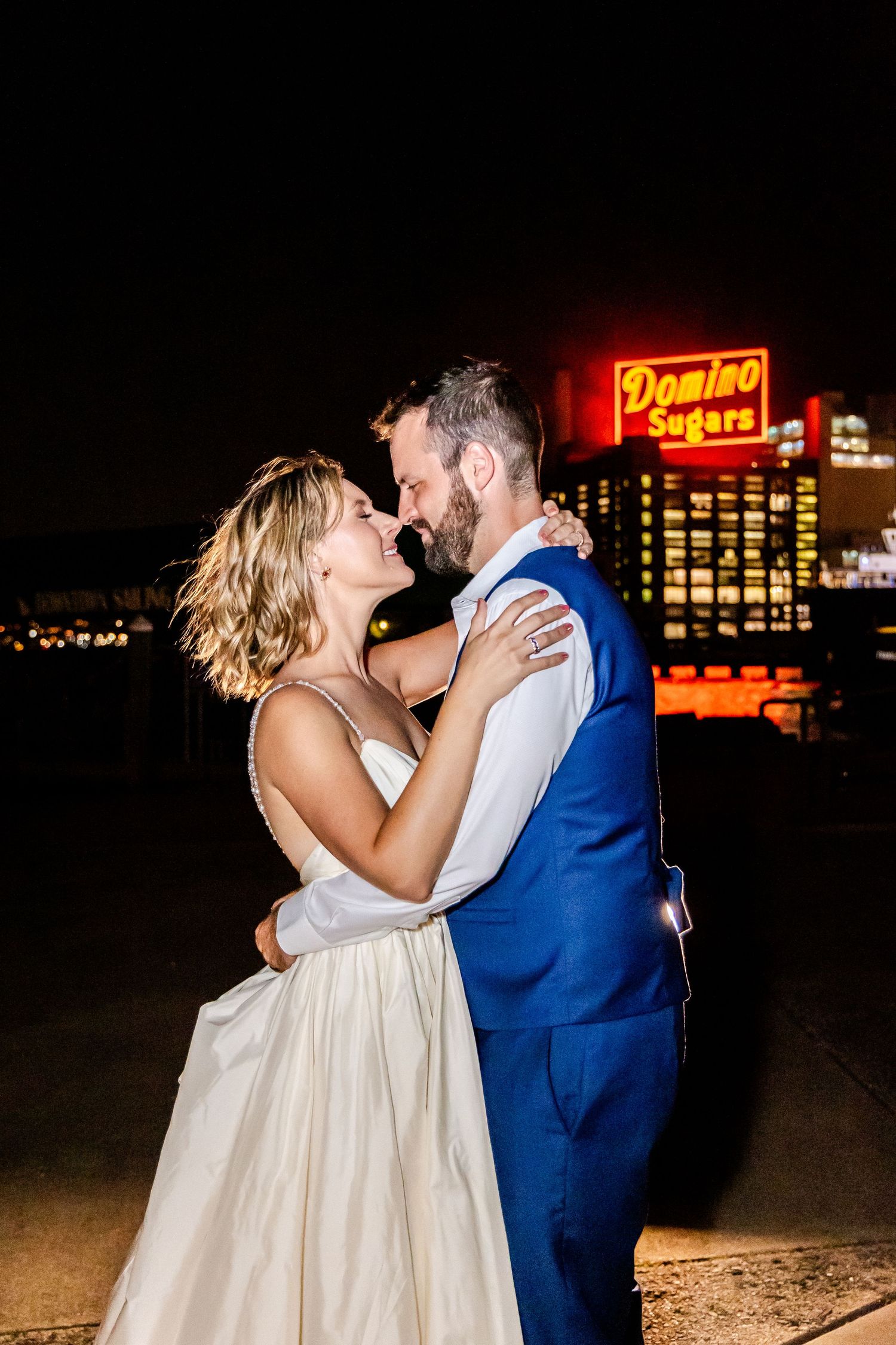 Romantic nighttime wedding photo with illuminated Domino Sugar sign in background.