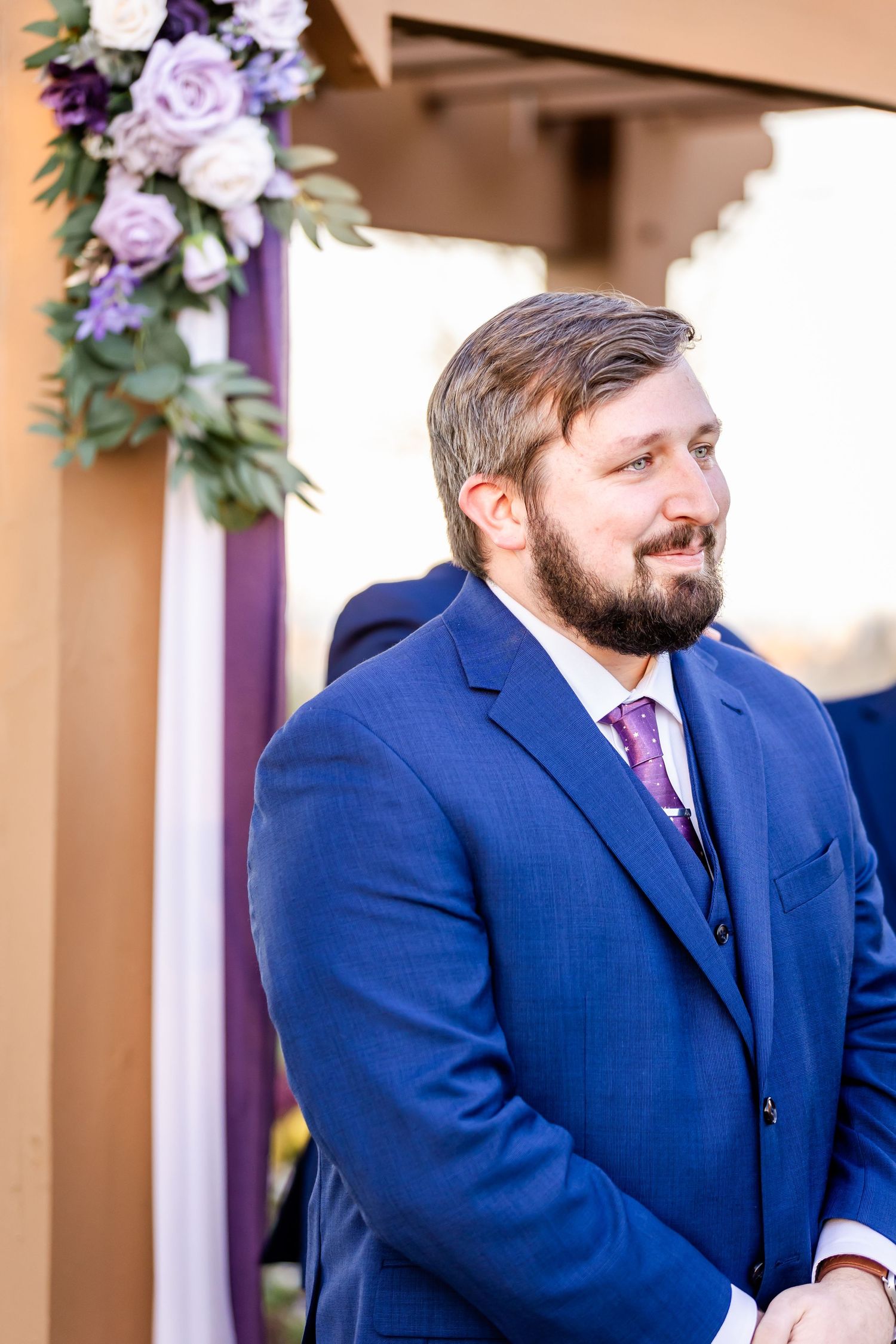 Groom in navy suit stands by floral arch decorated with purple and white flowers.