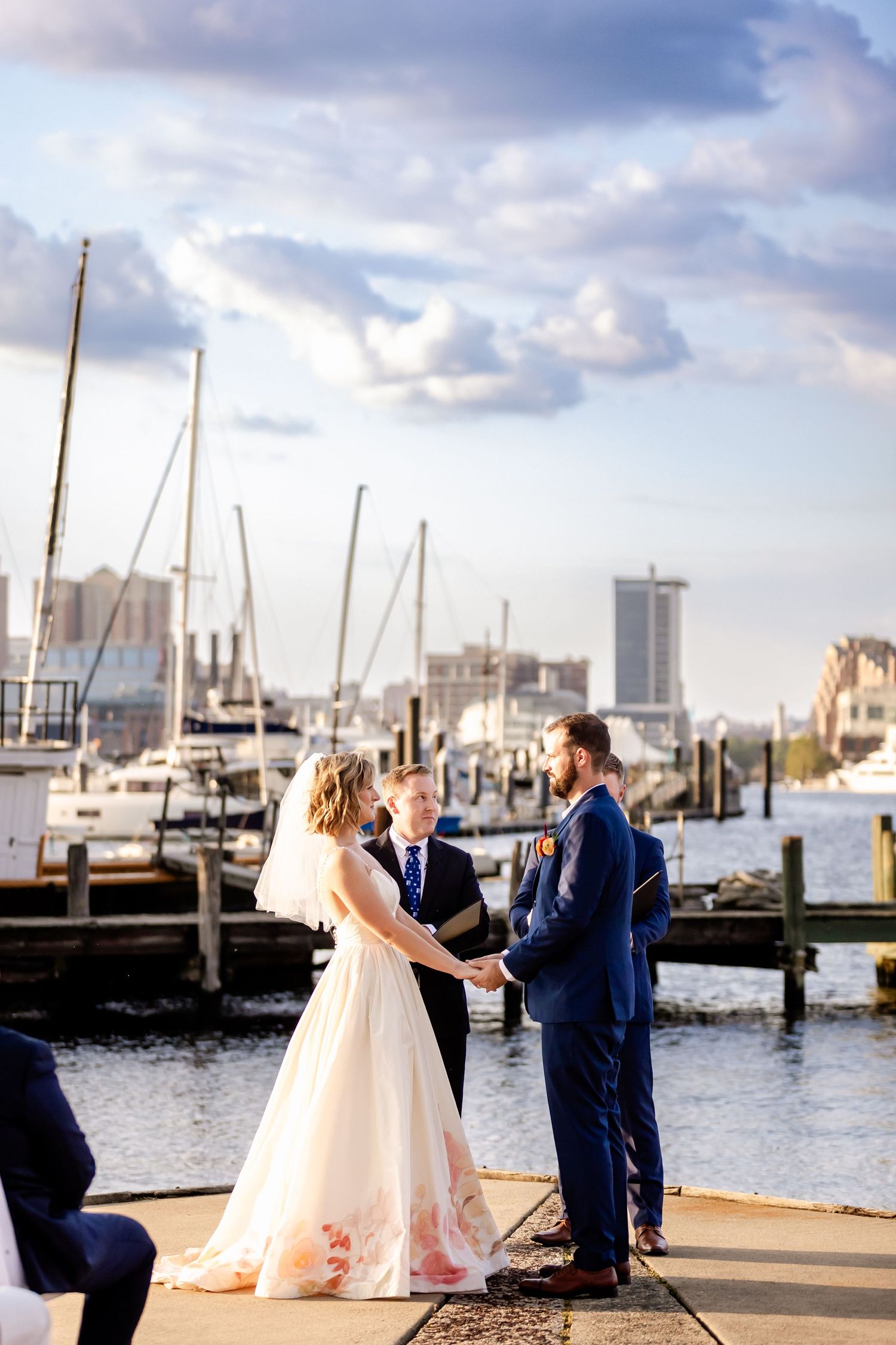 Couple exchanges vows during waterfront marina wedding ceremony with boats and cityscape in background.