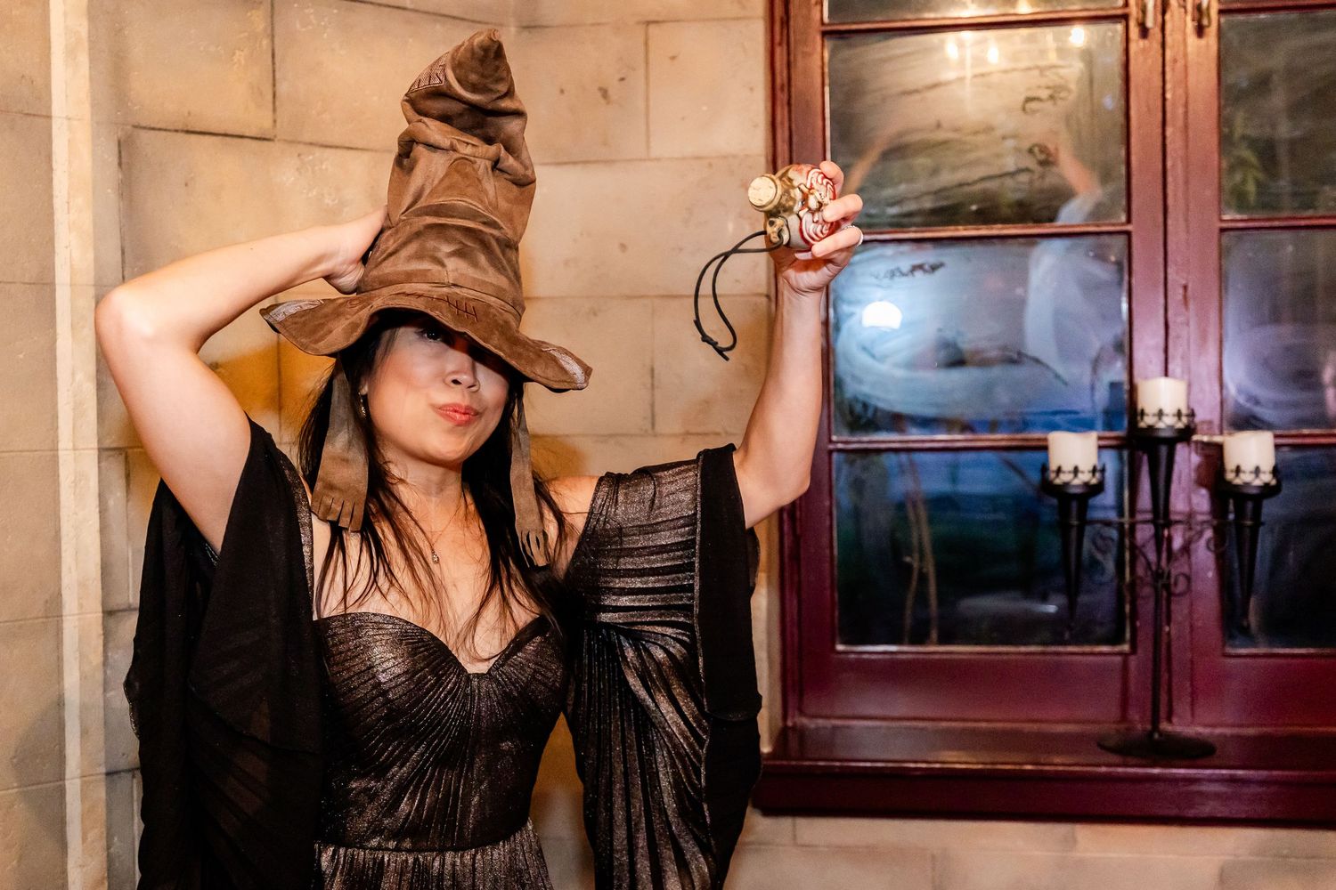 Person wearing a witch-style hat poses dramatically against a stone wall with vintage decor.