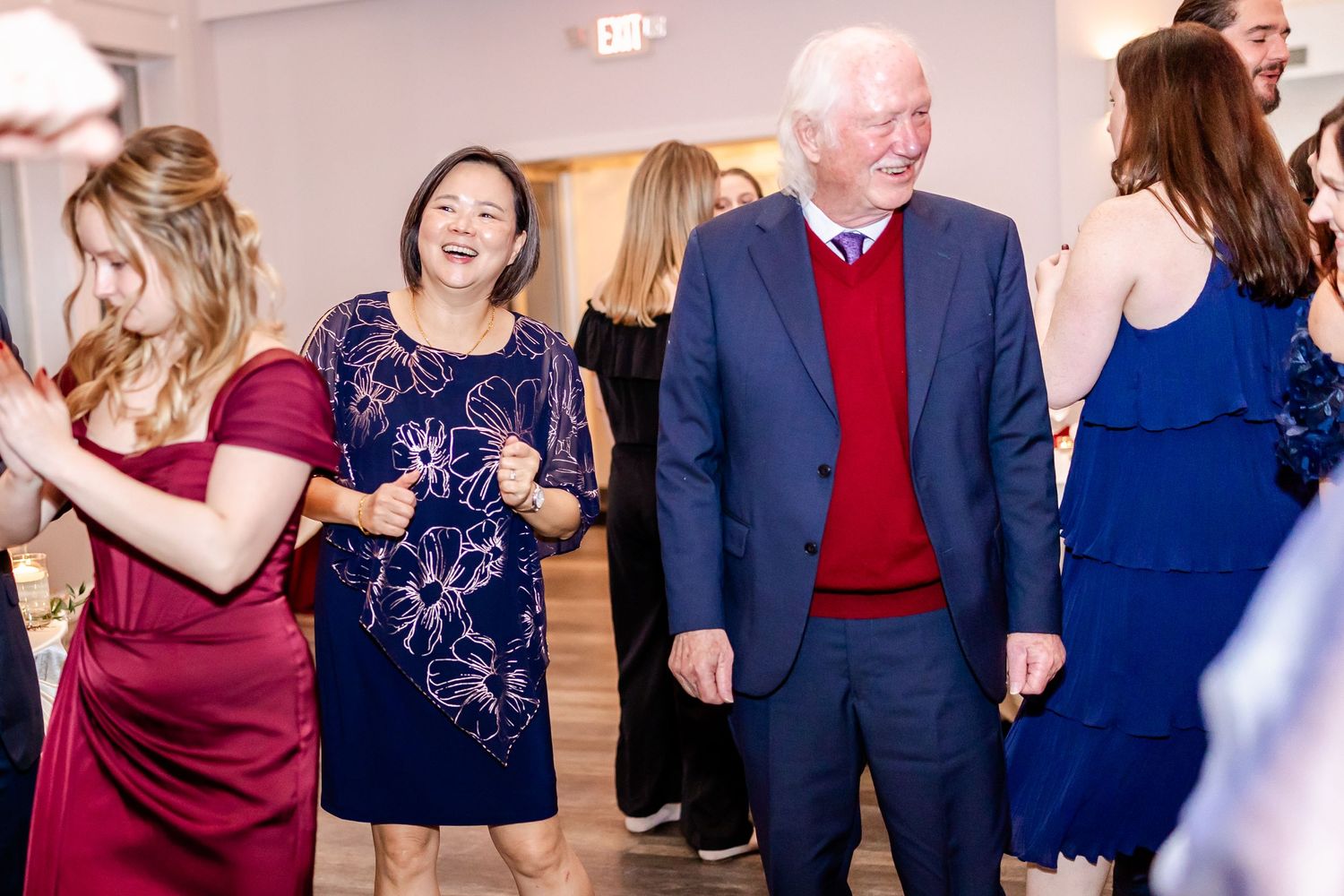 Wedding guests dance together in formal attire including navy blue suits and burgundy dresses at an indoor reception.