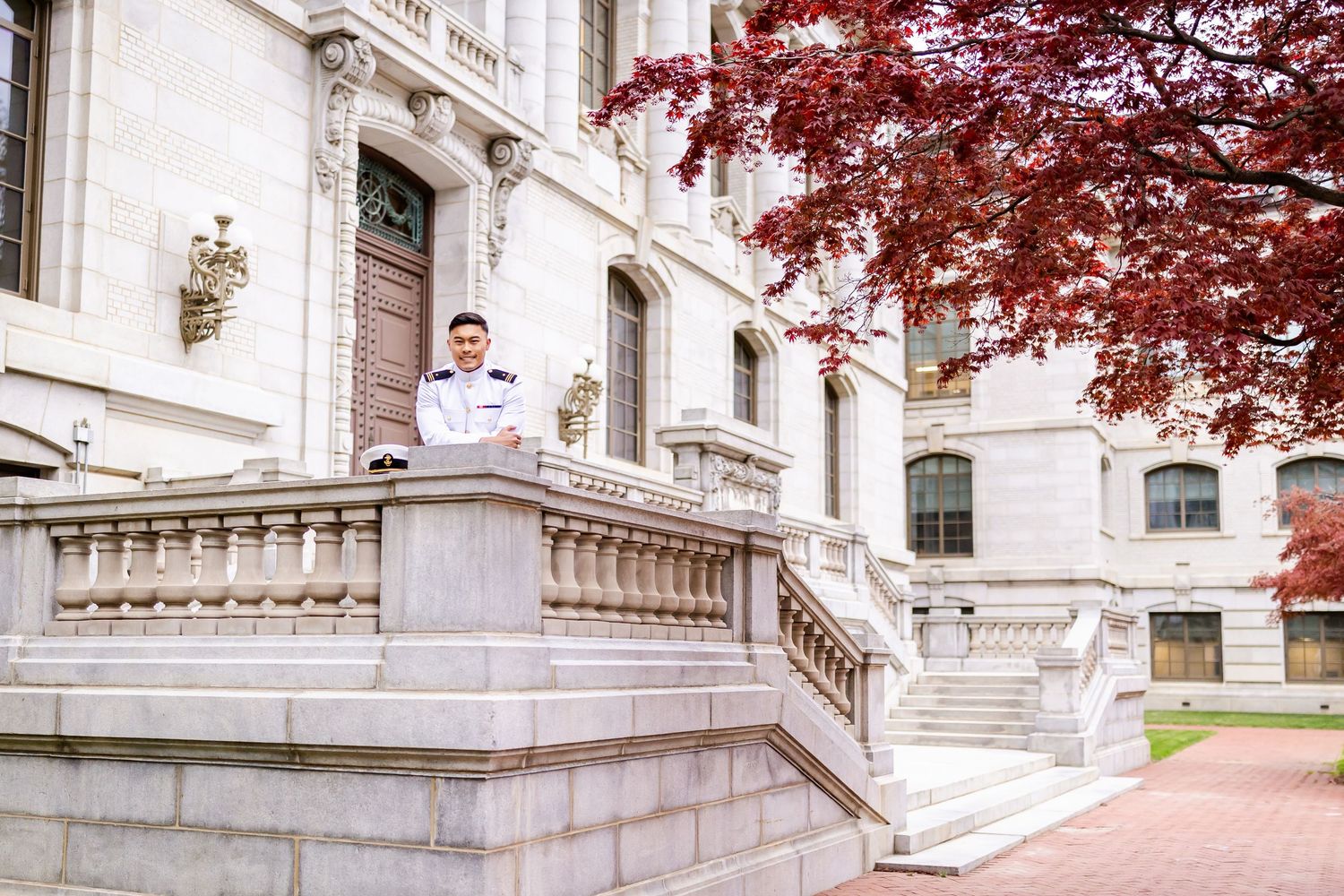 Naval officer in dress whites poses on ornate stone steps of a classical building with red autumn foliage.