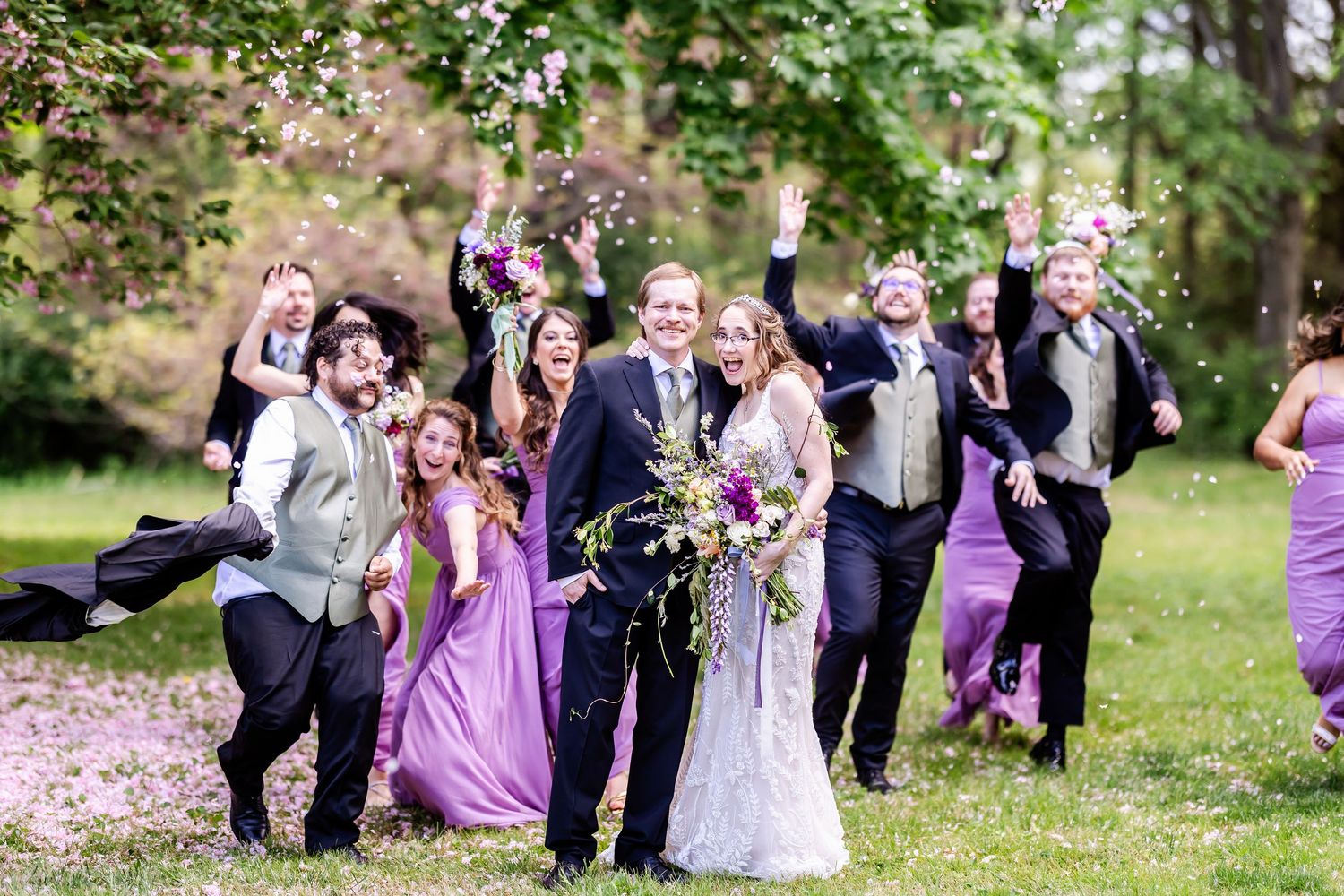 Wedding party in black tuxedos and purple bridesmaid dresses pose playfully outdoors among spring blossoms.