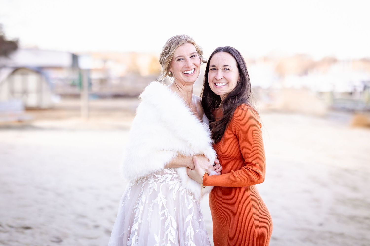 Two people embrace and laugh together outdoors, one wearing a white wedding dress and one in an orange dress.