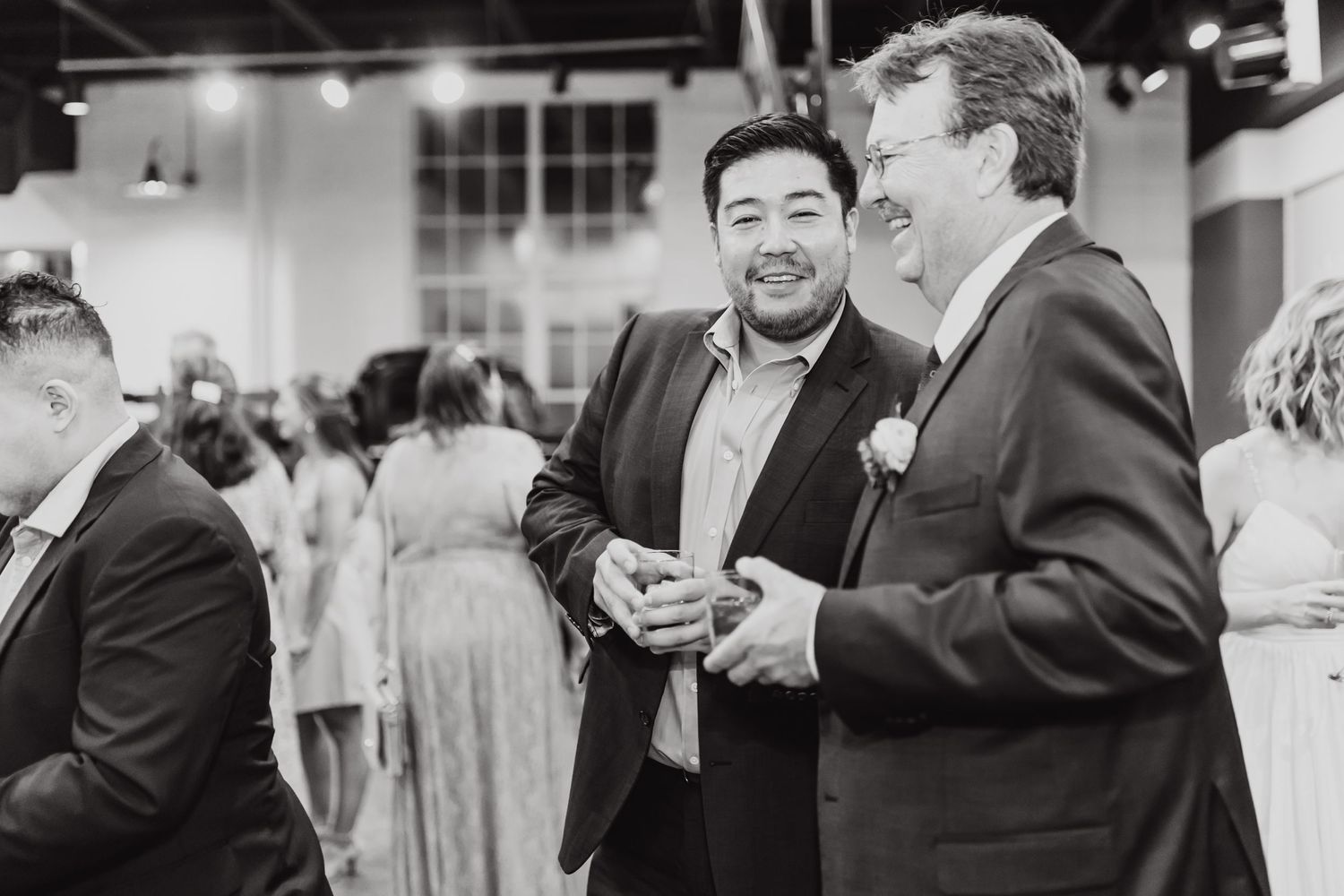 Wedding guests in formal attire share a candid moment at an indoor reception in black and white.