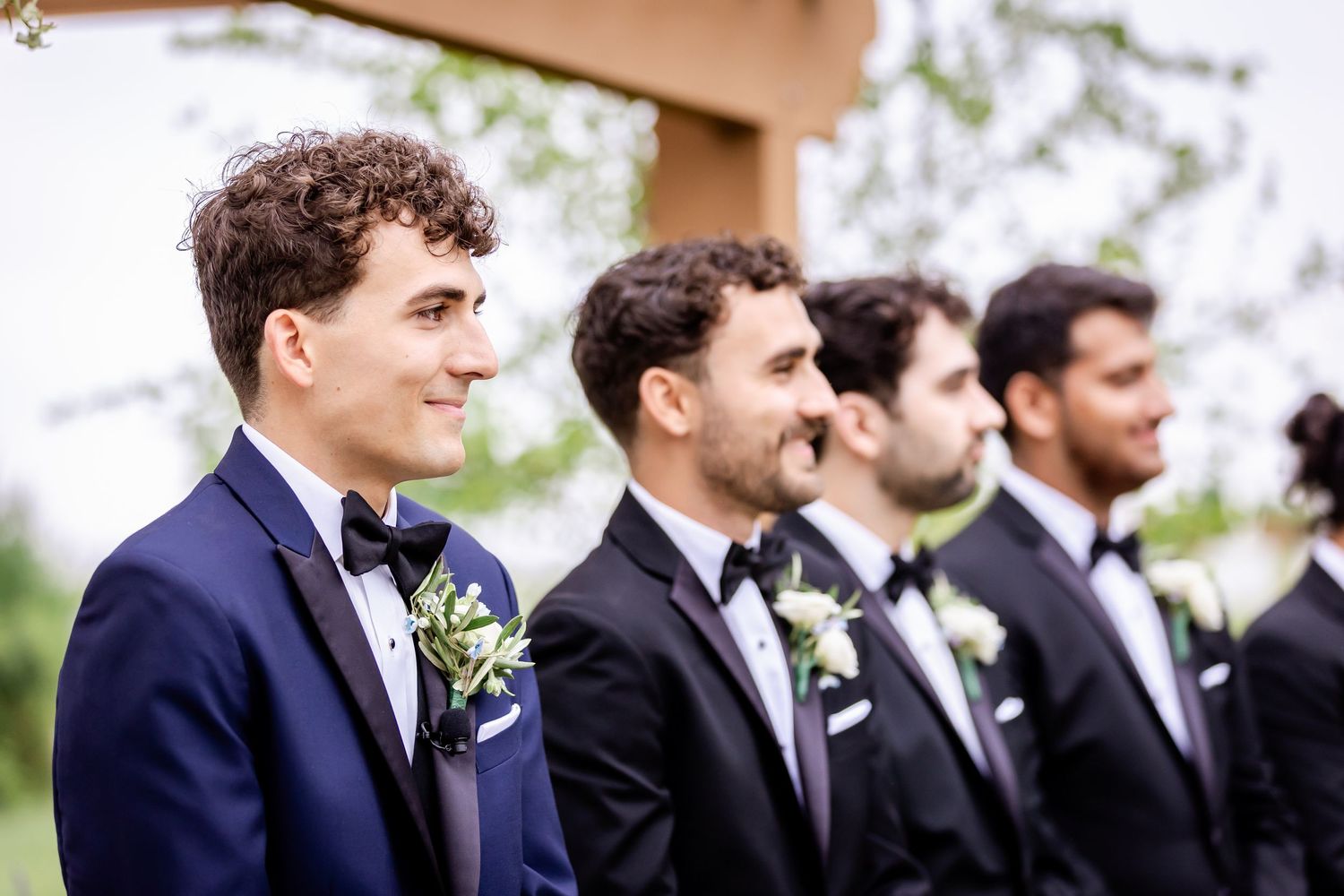 Groomsmen in navy suits and black bowties stand in line at an outdoor wedding ceremony.