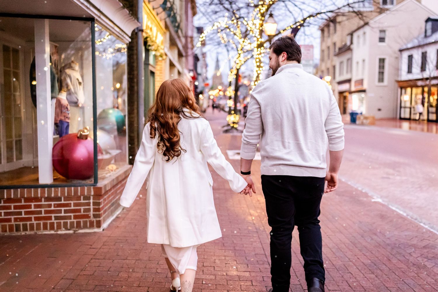A couple in white outfits walks hand in hand down a charming cobblestone street lined with shops and cafes.