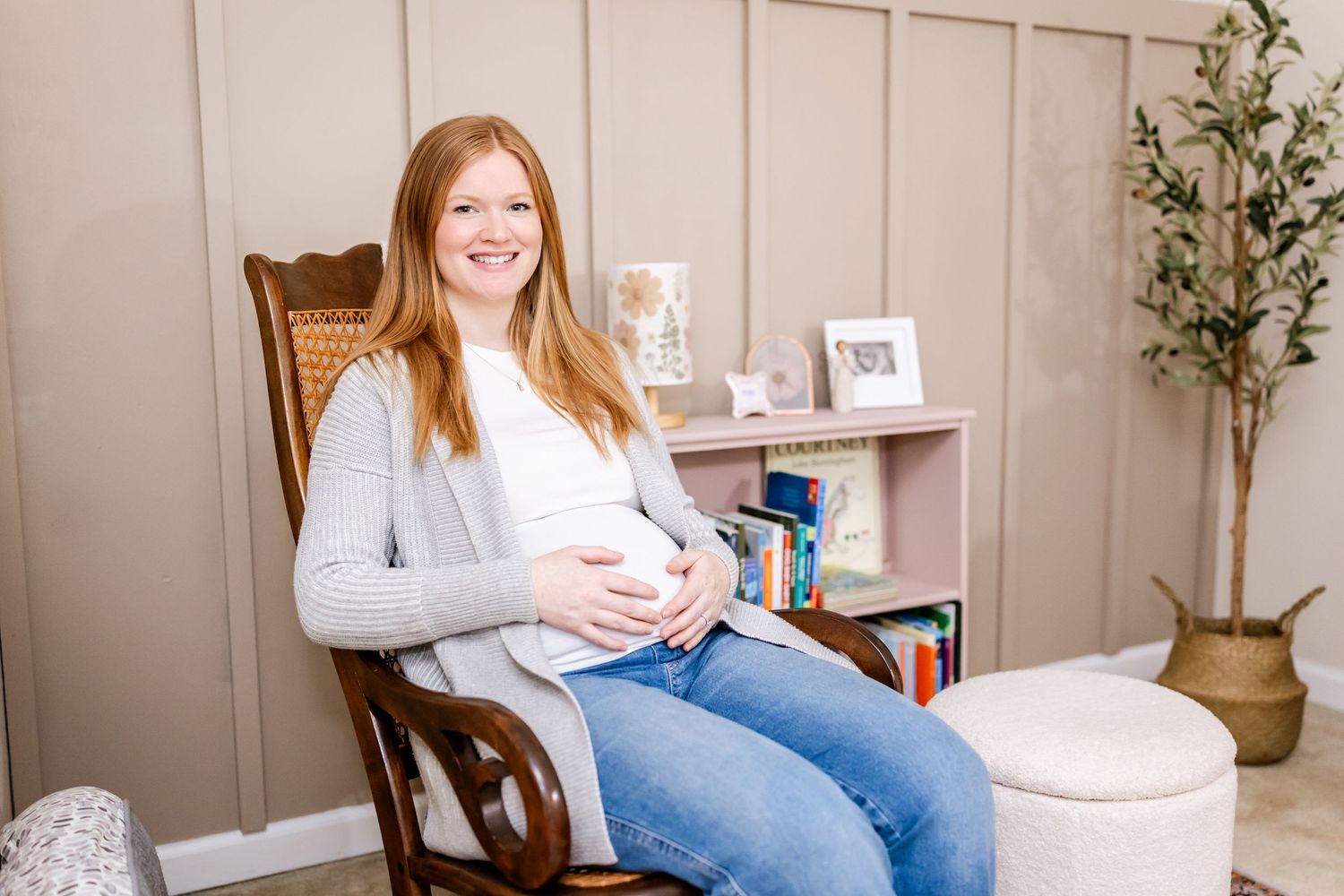 Person wearing light gray sweater and jeans sits in wooden chair next to bookshelf in cozy home setting.