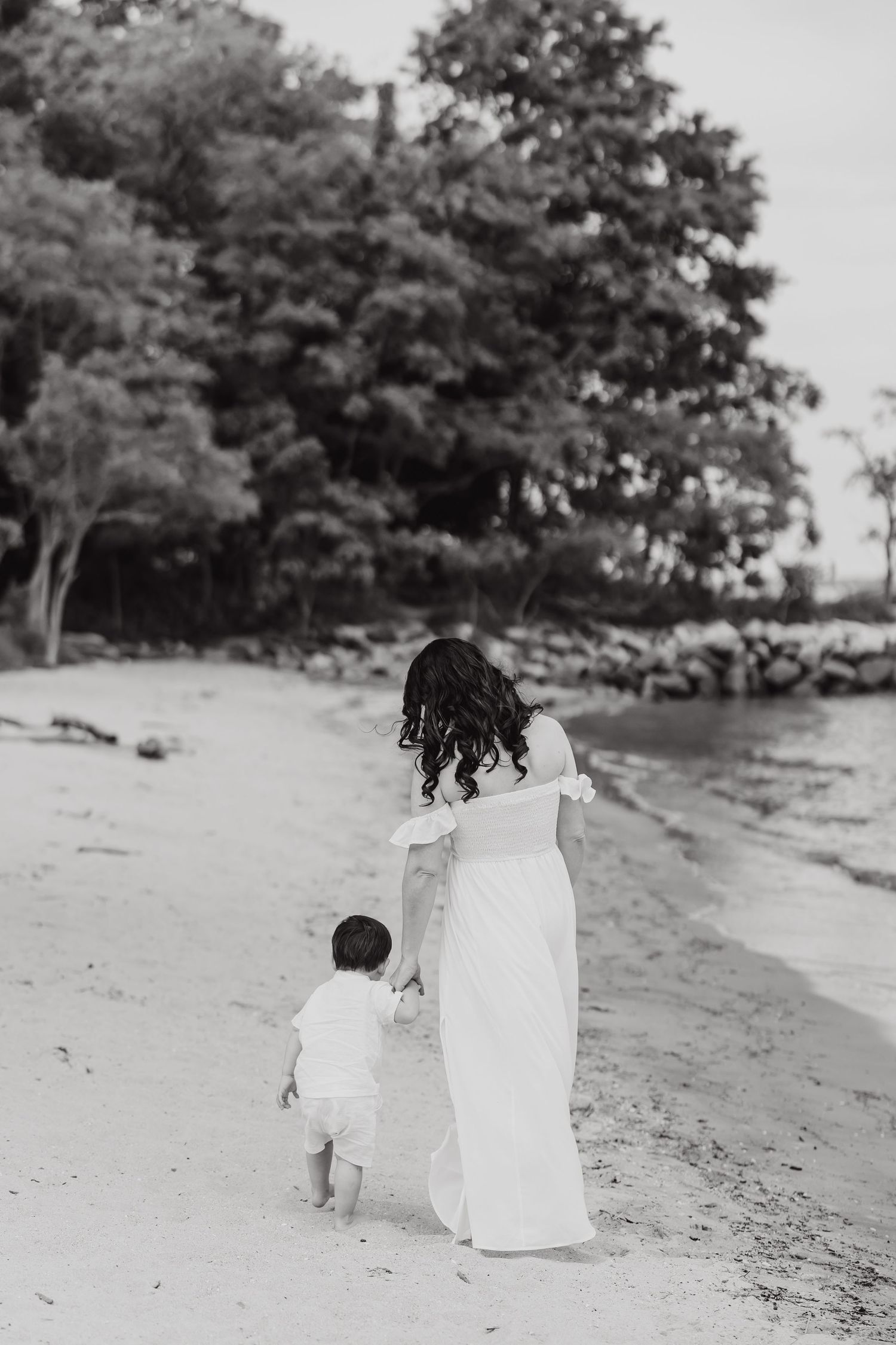 A black and white photo captures a tender moment between parent and child walking along a beach shoreline.